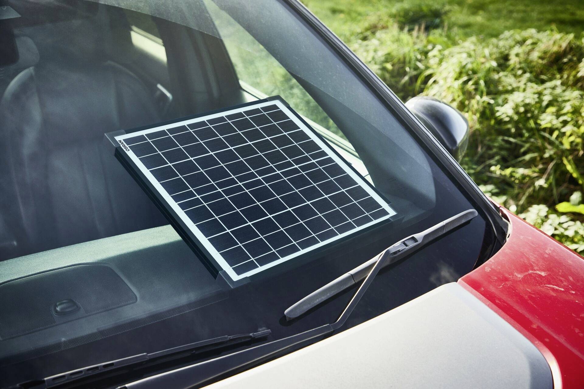 Ein Solarpanel liegt auf der Windschutzscheibe eines roten Autos, offenbar zum Laden der Batterie mithilfe von Sonnenenergie.