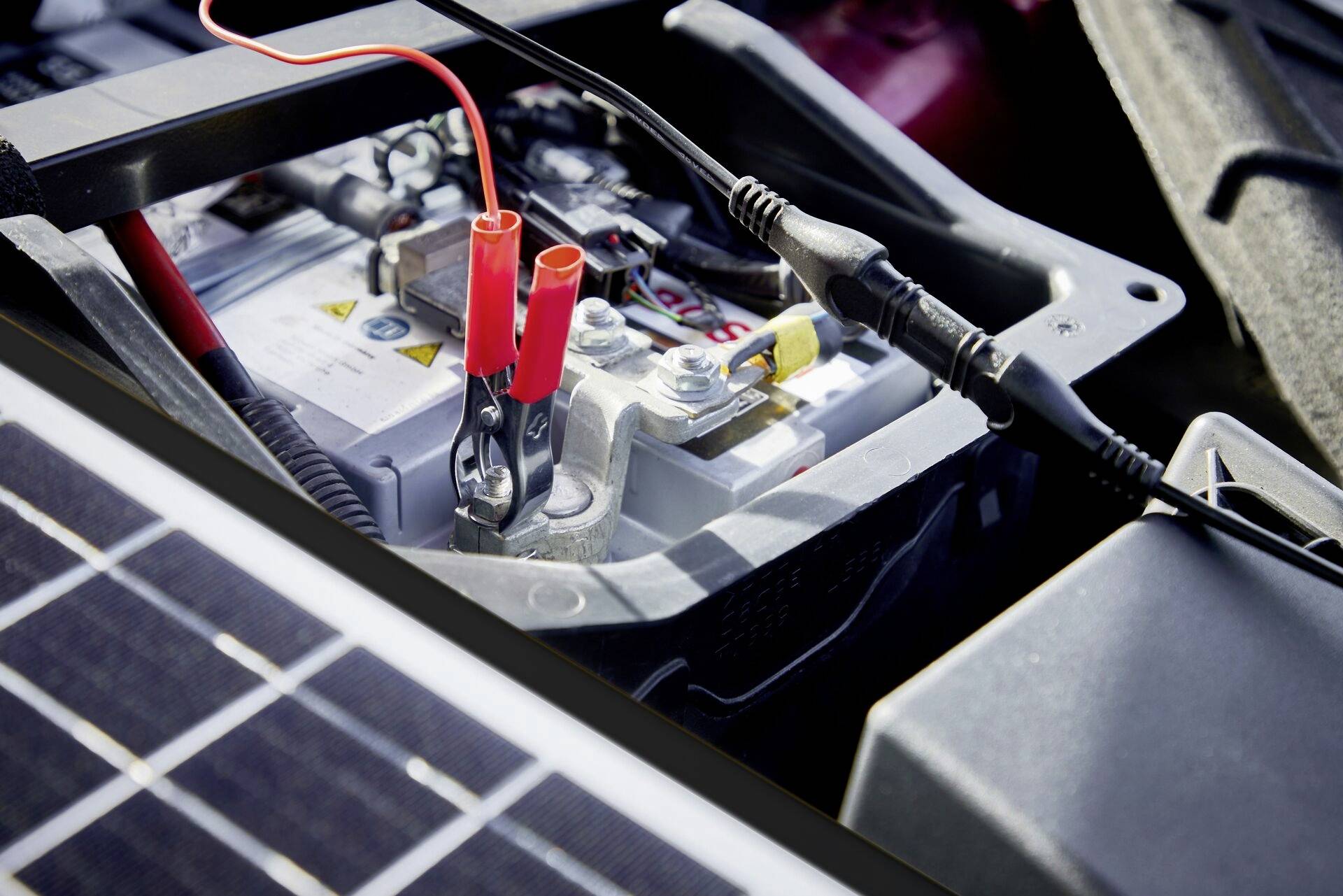 Autospringer mit Starthilfekabeln, die an eine Autobatterie angeschlossen sind. Im Hintergrund sind Motorteile zu sehen.