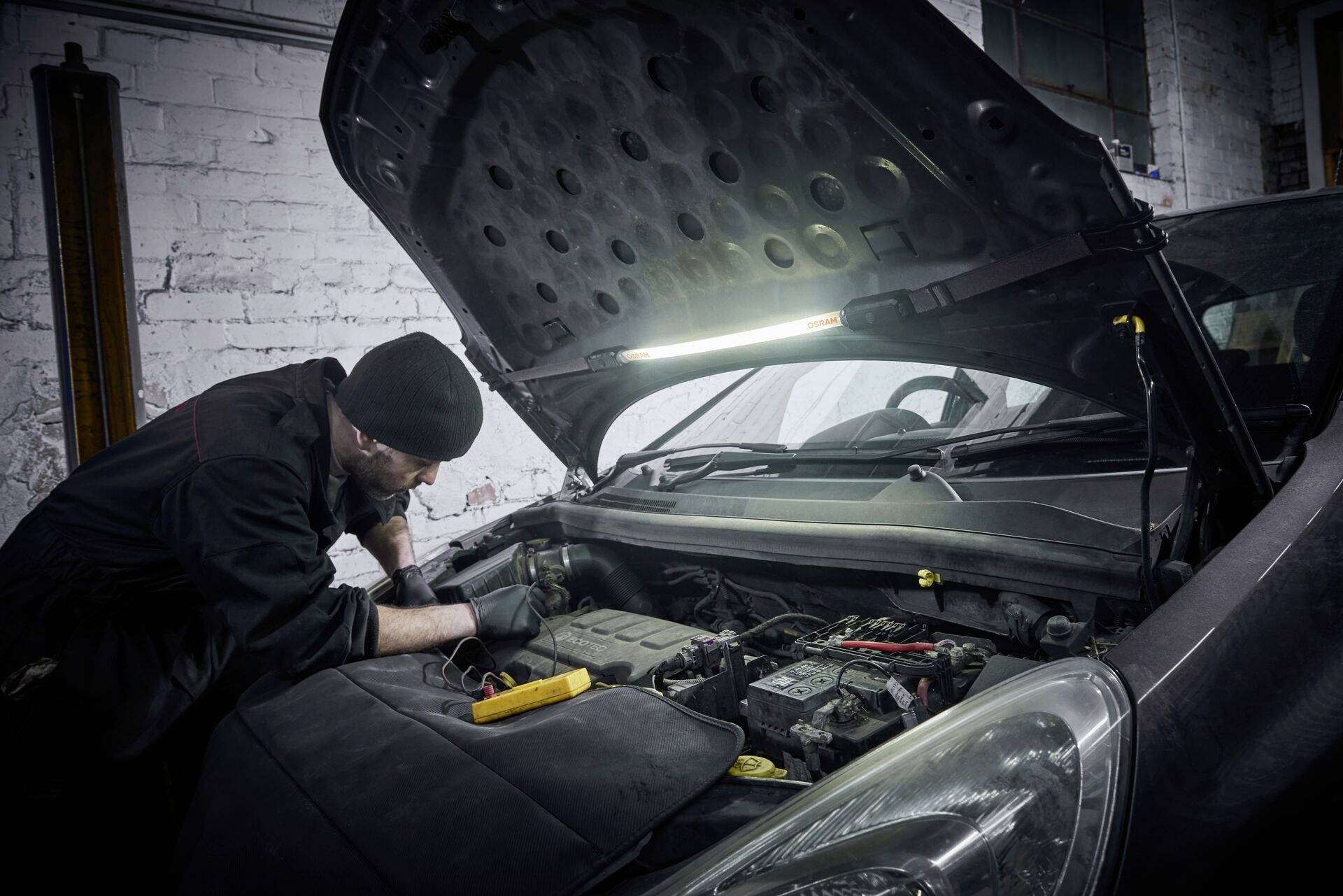 Mechaniker in einer Werkstatt, arbeitet unter der geöffneten Motorhaube eines Autos. Der Raum ist spärlich beleuchtet.