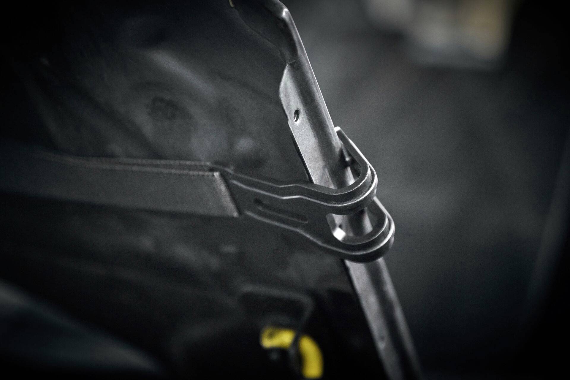 Nahaufnahme eines schwarzen Metallhakens, der einen Gurt hält, vermutlich an einer Autotür befestigt. Der Hintergrund ist unscharf.