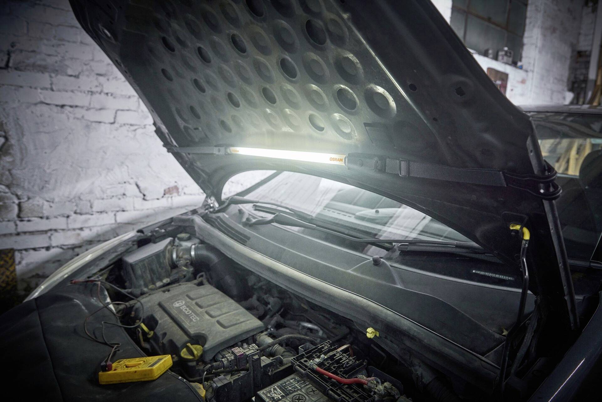 Ein Auto steht mit geöffneter Motorhaube in einer Werkstatt. Ein Diagnosegerät liegt neben dem Motor, um Wartungsarbeiten zu unterstützen.