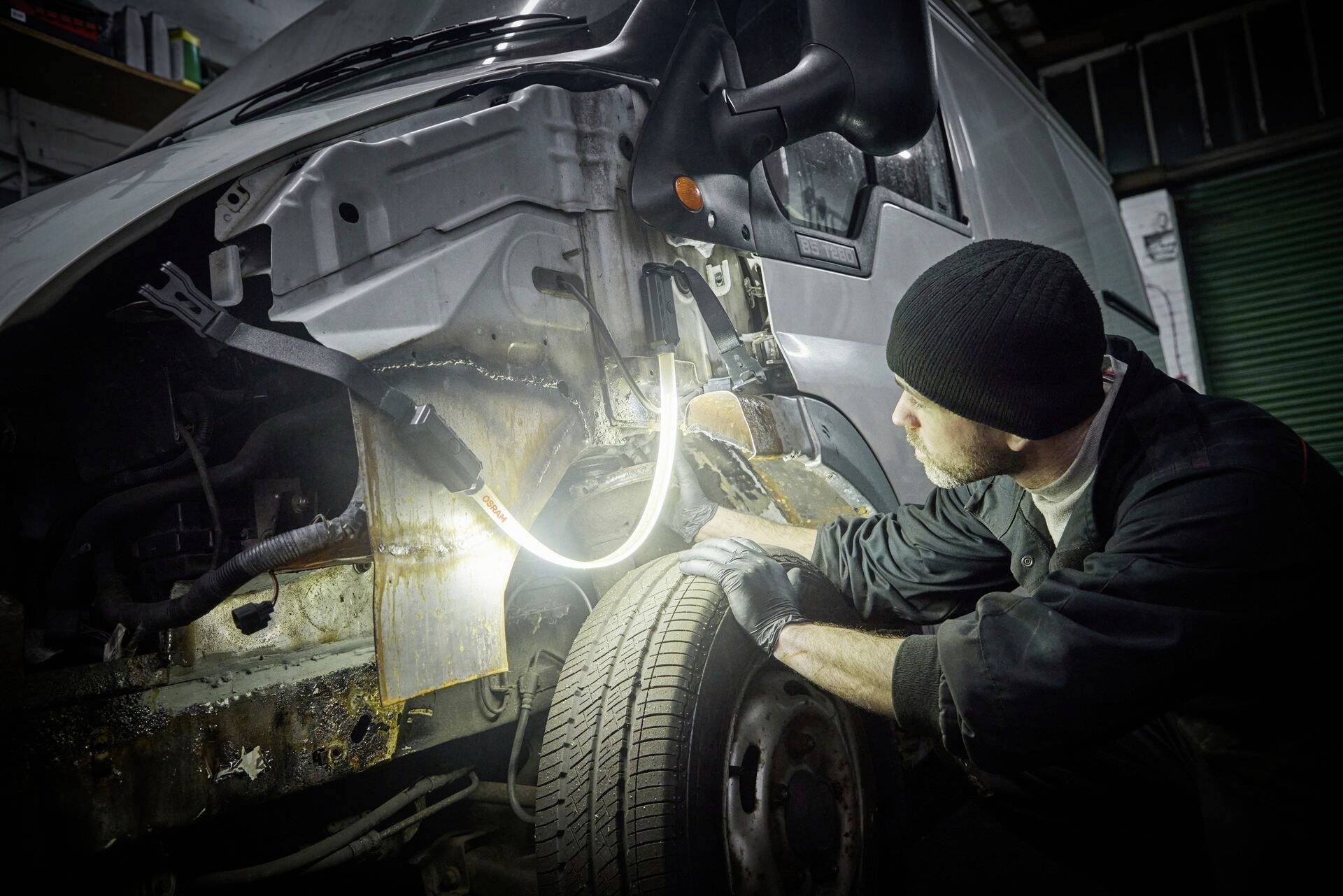 Mechaniker inspiziert beschädigten Kotflügel eines Transporters in einer Werkstatt. Beleuchtung zeigt deutliche Rostspuren am Fahrzeugrahmen.