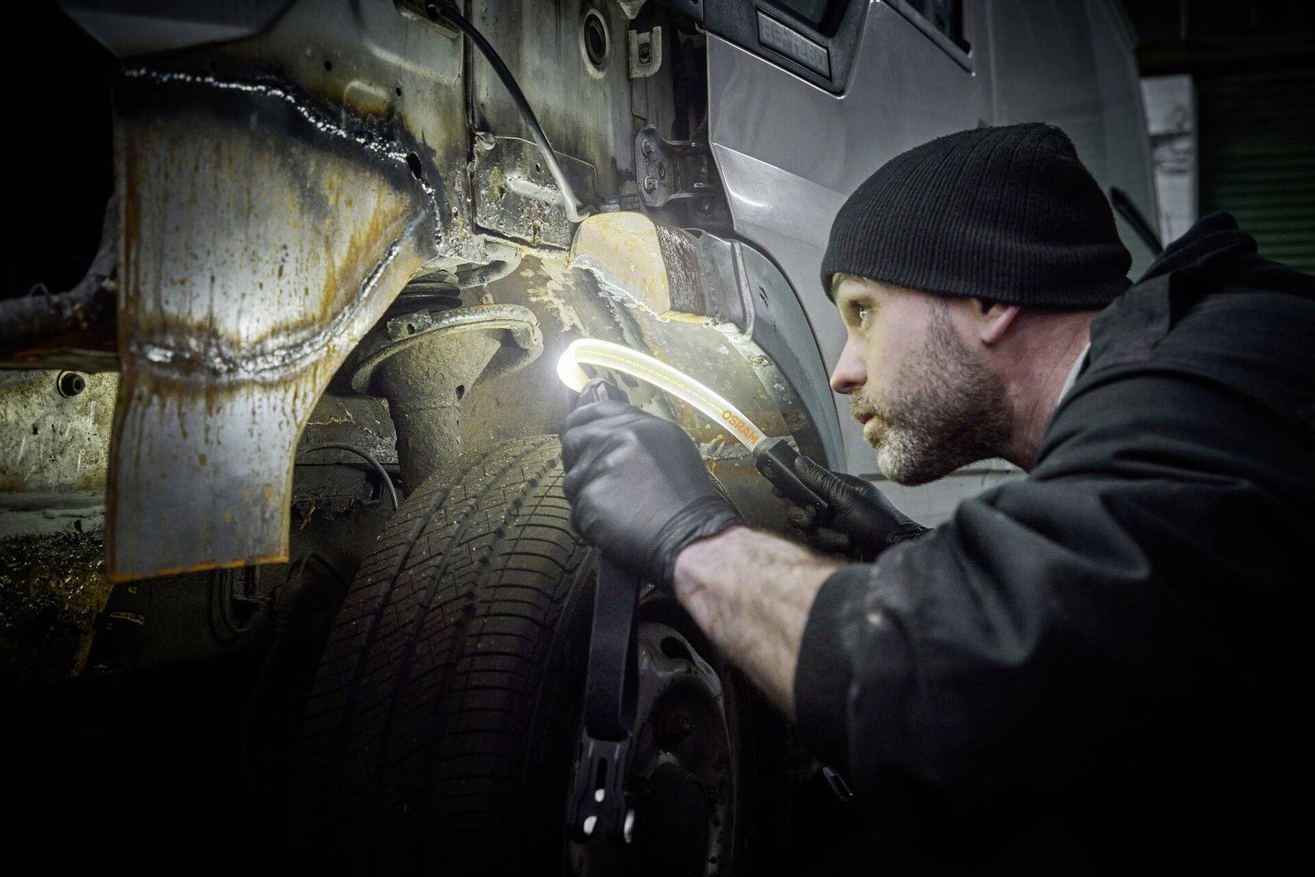 Ein Mechaniker inspiziert sorgfältig den rostigen Radkasten eines Fahrzeugs in einer Werkstatt. Er nutzt eine Leuchte für bessere Sicht.