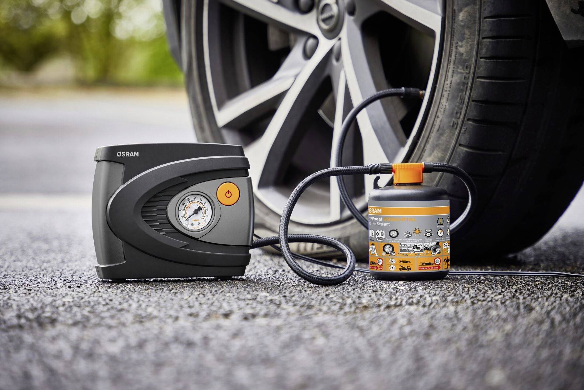 Tragbarer Reifenkompressor neben einem Auto, bereit zum Aufpumpen. Gerät mit Druckanzeige und Schlauch, auf Asphalt platziert.