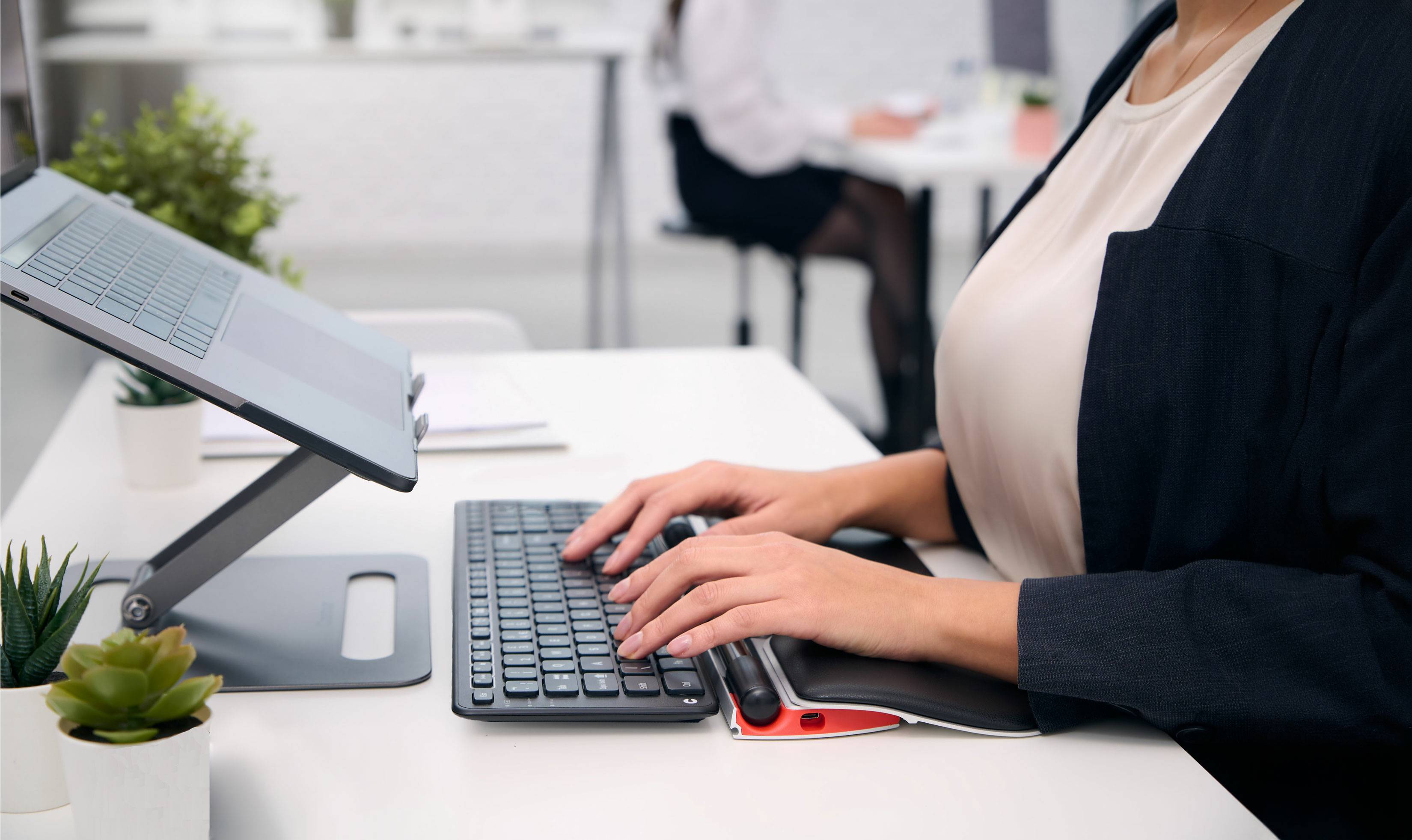 Eine Person arbeitet an einem höhenverstellbaren Laptopständer an einem modernen Büroschreibtisch. Im Hintergrund ist ein weiteres Büro zu sehen.