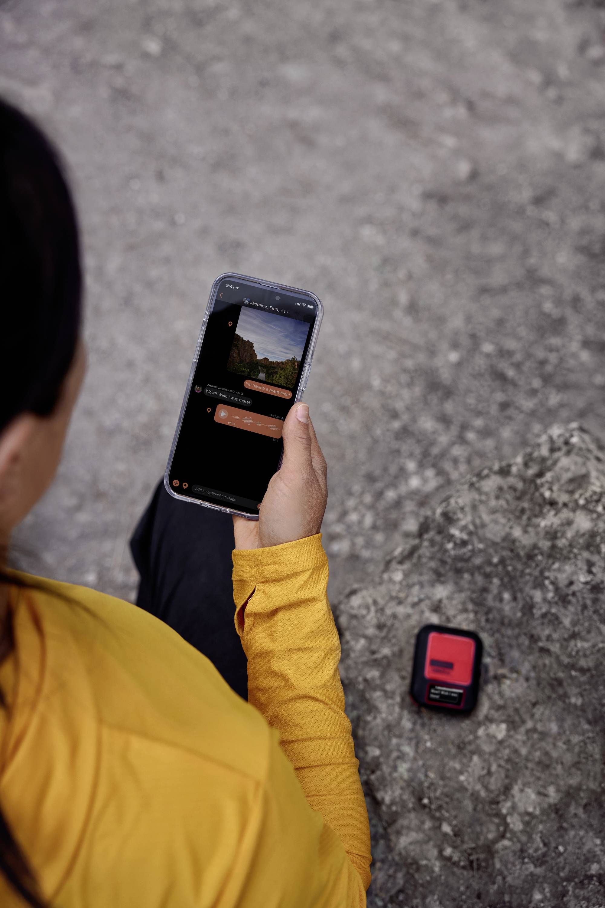 Eine Person in gelber Jacke schaut auf ein Smartphone, während sie draußen auf einem felsigen Untergrund sitzt. Ein kleines rotes Gerät liegt neben ihr.