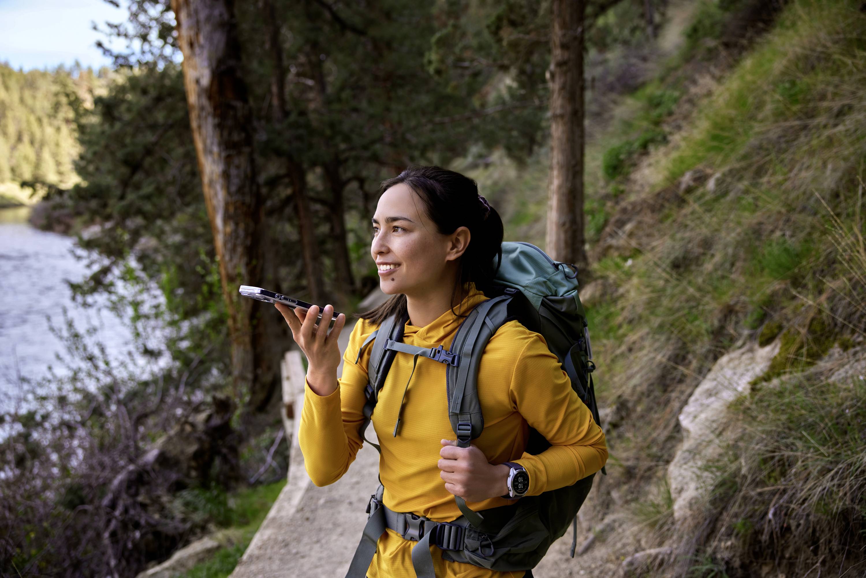 Frau auf Wanderweg spricht in ihr Smartphone. Sie trägt einen Rucksack und ist von Wald umgeben. Im Hintergrund ein Fluss.