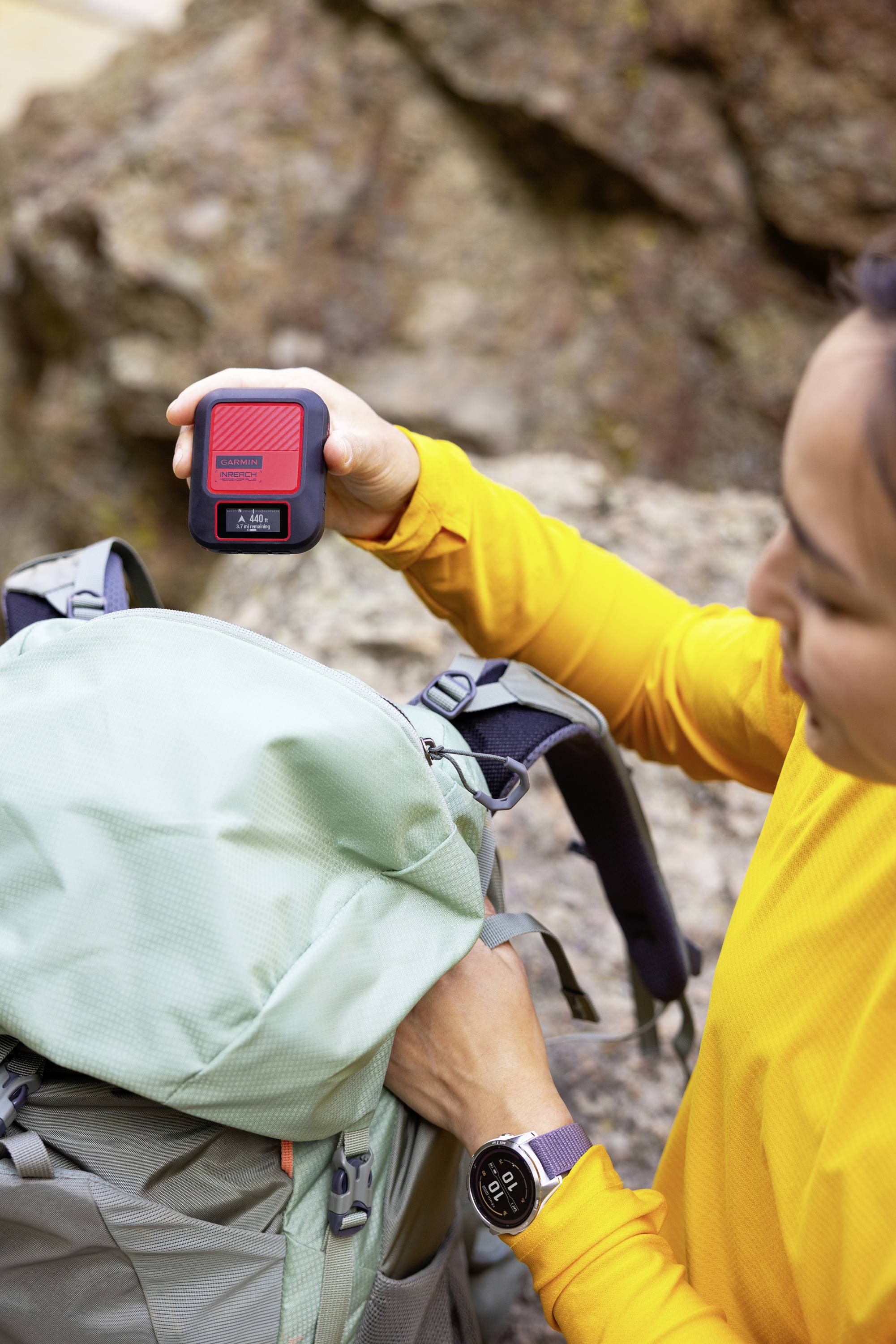Eine Person in gelbem Oberteil hält einen kleinen roten Gegenstand, während sie einen Rucksack öffnet. Im Hintergrund Felsen.
