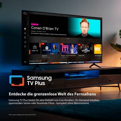 Ein Fernseher zeigt eine Streaming-Service-Oberfläche mit einer Show mit "Conan O'Brien". Das Logo liest sich "Samsung TV Plus". Ein Tisch und eine Lampe sind sichtbar.