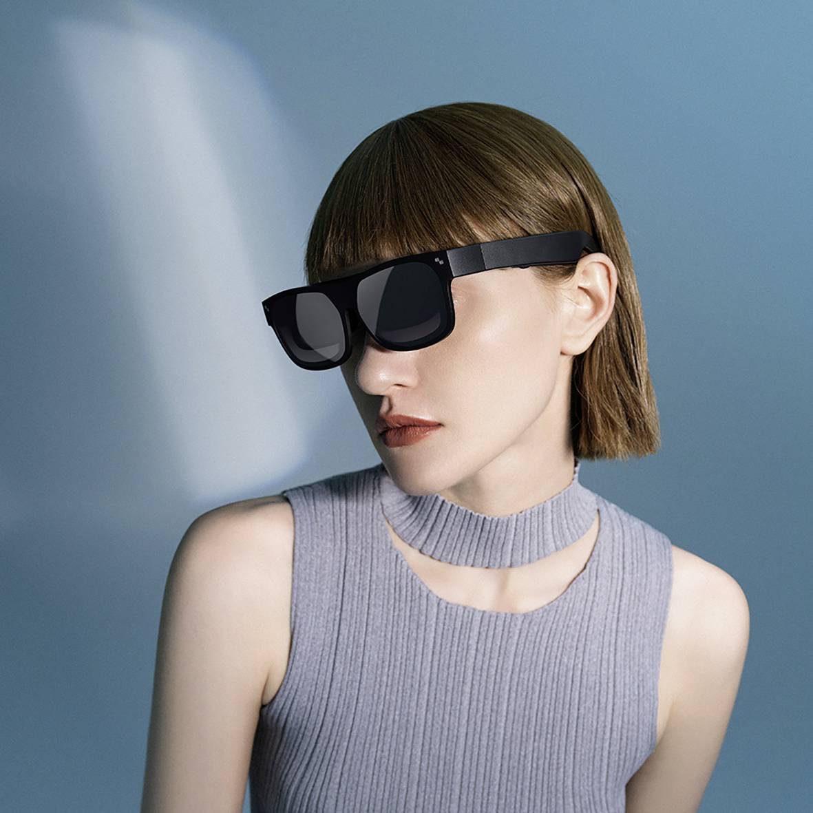 Eine Person mit glattem Haar trägt eine futuristische, schwarze VR-Brille und ein graues, ärmelloses Oberteil, sitzt in einem neutralen Raum.