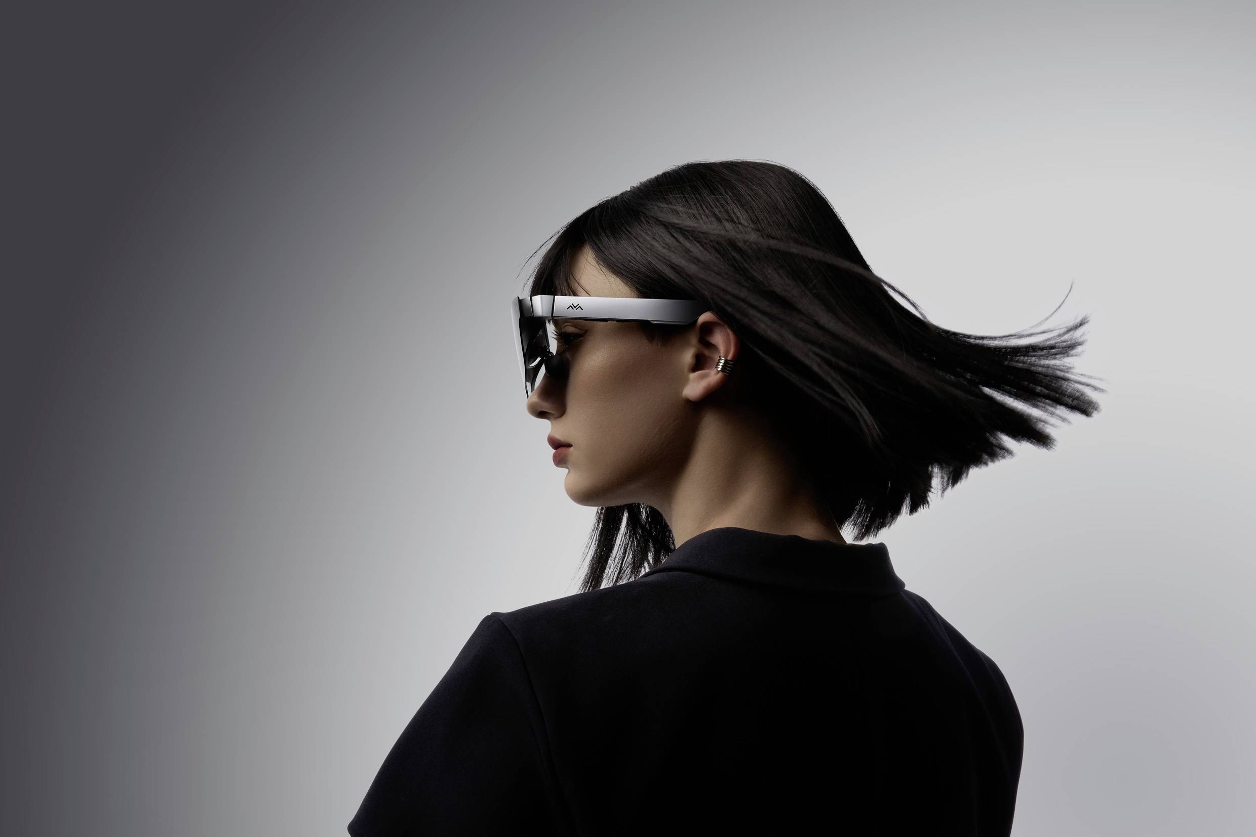 Frau mit dunklem Haar trägt eine moderne, weiße Datenbrille. Ihr Haar weht im Wind, Hintergrund ist minimalistisch grau.