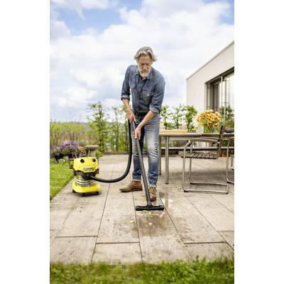 Ein Mann in Jeans und Hemd saugt die Holzterrasse mit einem gelben Staubsauger ab. Im Hintergrund ein Tisch mit Blumen.