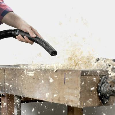 Eine Hand hält einen Staubsaugerschlauch über einer Werkbank, auf der Holzspäne verstreut sind, während ein Hobel rechts sichtbar ist.