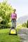 Eine Frau mäht den Rasen mit einem gelben Elektro-Rasenmäher in einem Garten, umgeben von Bäumen und Bergen im Hintergrund.