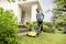 Ein Mann mäht den Rasen mit einem gelben Rasenmäher vor einem weißen Haus. Umgebung zeigt blühende Pflanzen und grünes Laub.