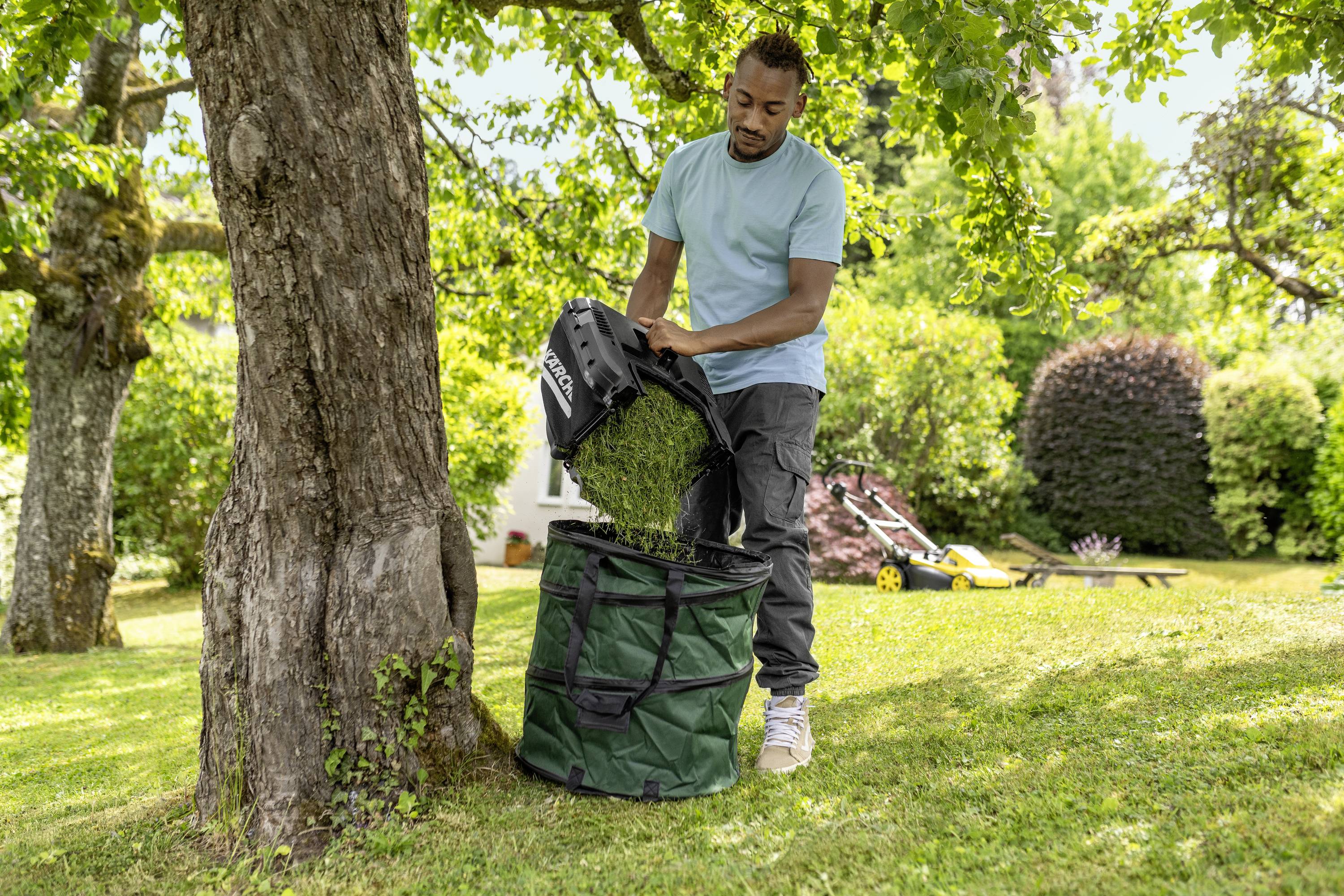 Eine Person leert den Grasfangkorb eines Rasenmähers in einen grünen Gartensack unter einem Baum im Garten.