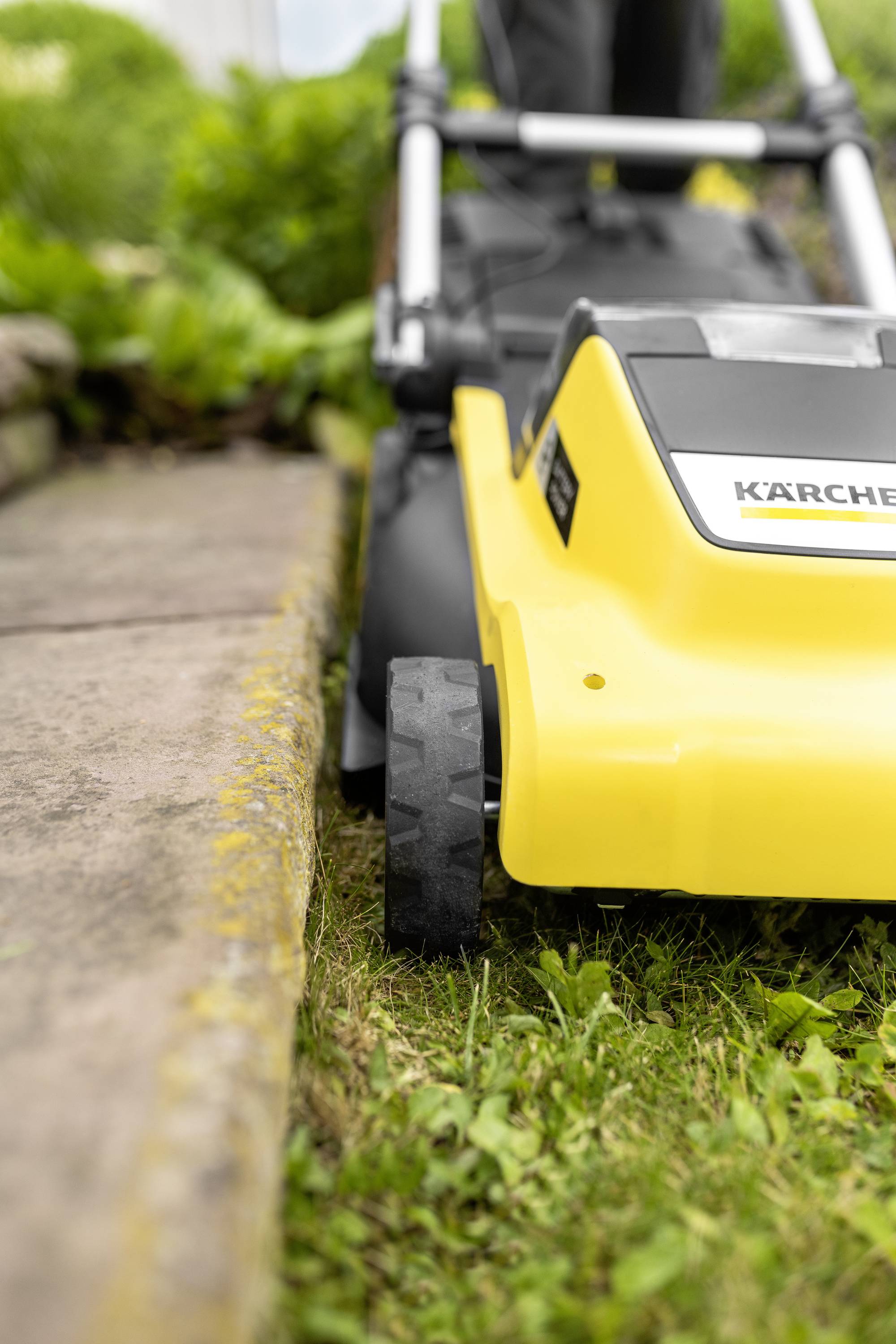 Rasenmäher schneidet Gras entlang eines Gehwegs in einem Garten. Fokus auf das gelbe Gerät und seine Räder. Grünen Pflanzen im Hintergrund.