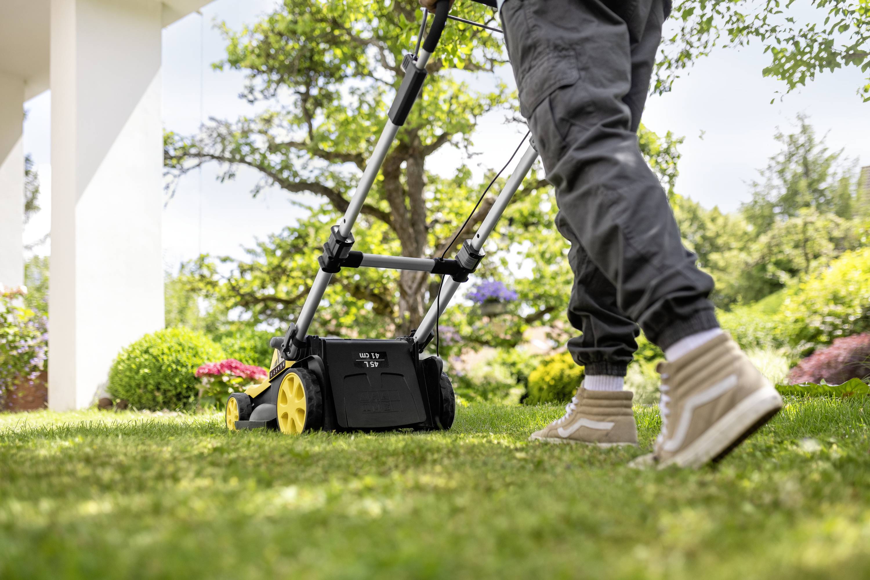 Eine Person mäht den Rasen mit einem Rasenmäher im Garten. Der Fokus liegt auf den Füßen der Person und dem Gerät.