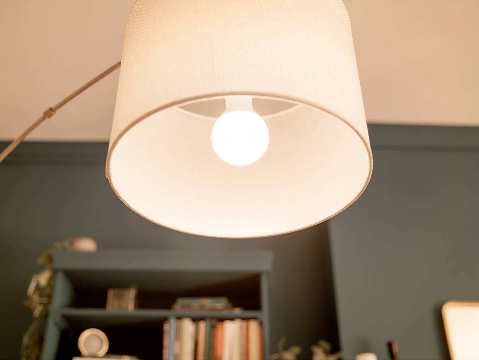 Stehlampe mit cremefarbenem Lampenschirm, darunter ein Regal mit Büchern und Dekor gegen eine petrolblaue Wand.