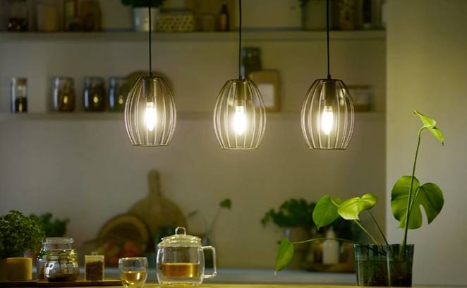 Drei hängende Lampen leuchten über einem Küchentresen. Darunter Pflanzen und Geschirr. Hintergrund mit unscharfen Küchenregalen.