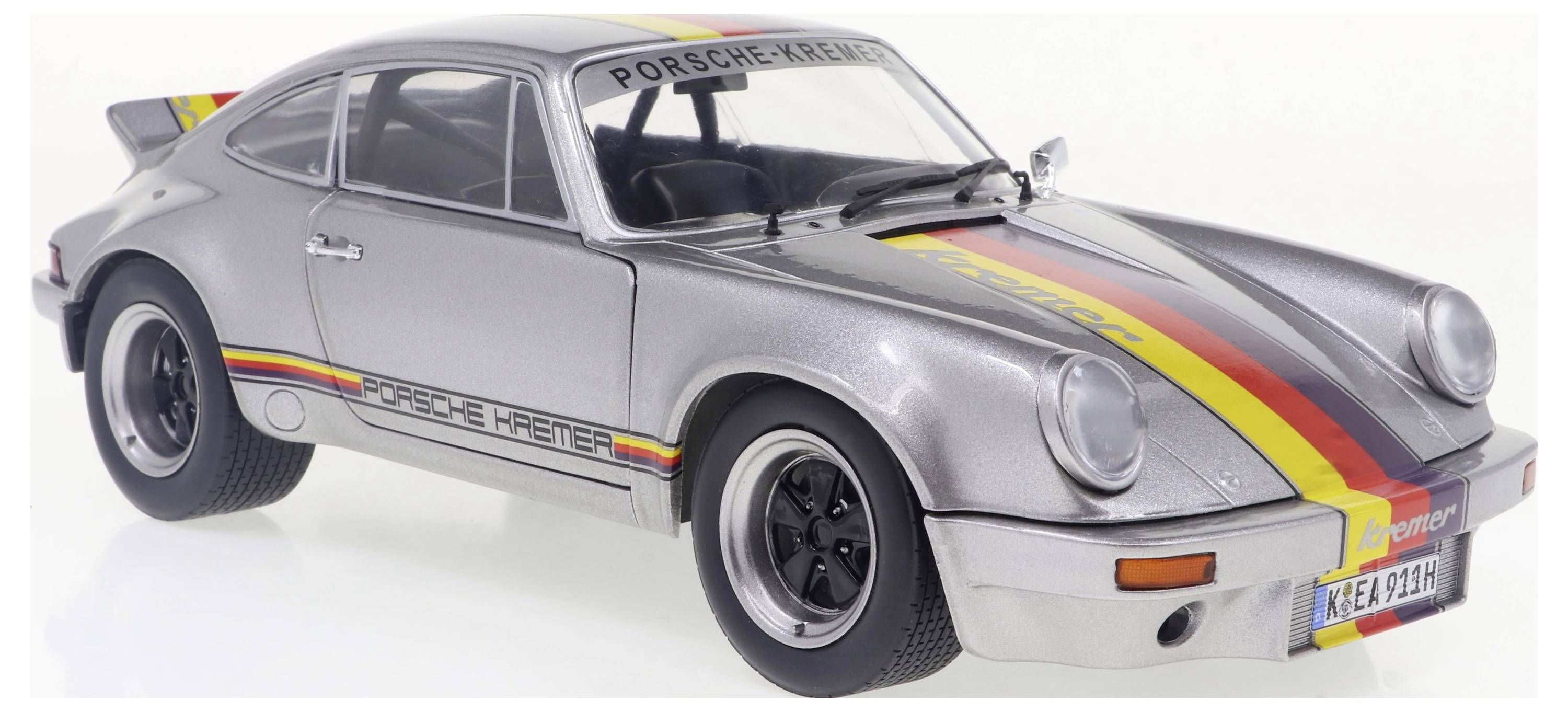 Silberner Sportwagen mit schwarz-rot-gelben Streifen und der Aufschrift 'Porsche Kremer' auf der Windschutzscheibe, von vorne rechts fotografiert.