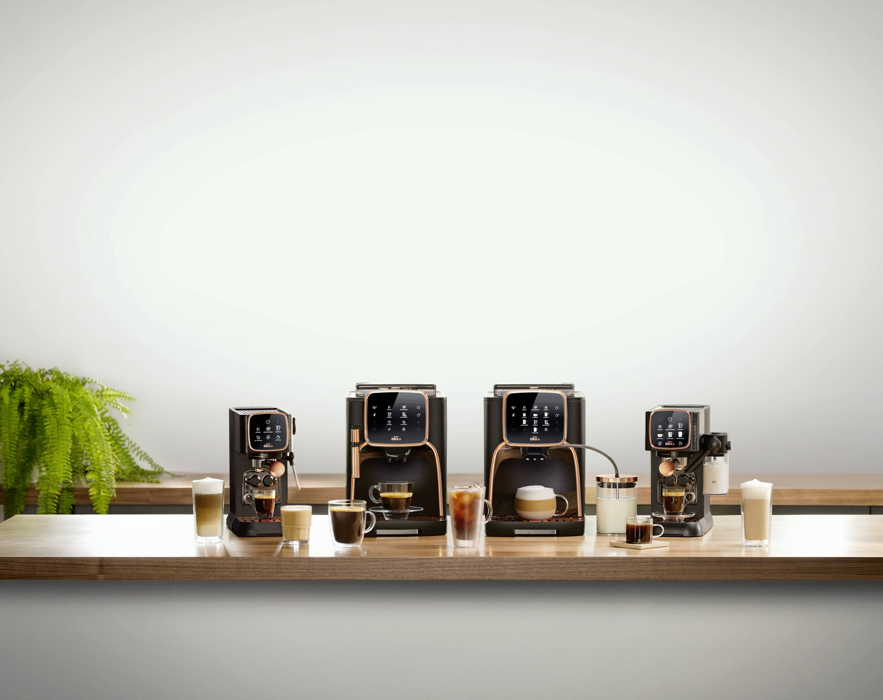 Drei moderne Kaffeemaschinen in einer Reihe auf einem Tisch, mit verschiedenen zubereiteten Kaffeegetränken und einer Topfpflanze links.