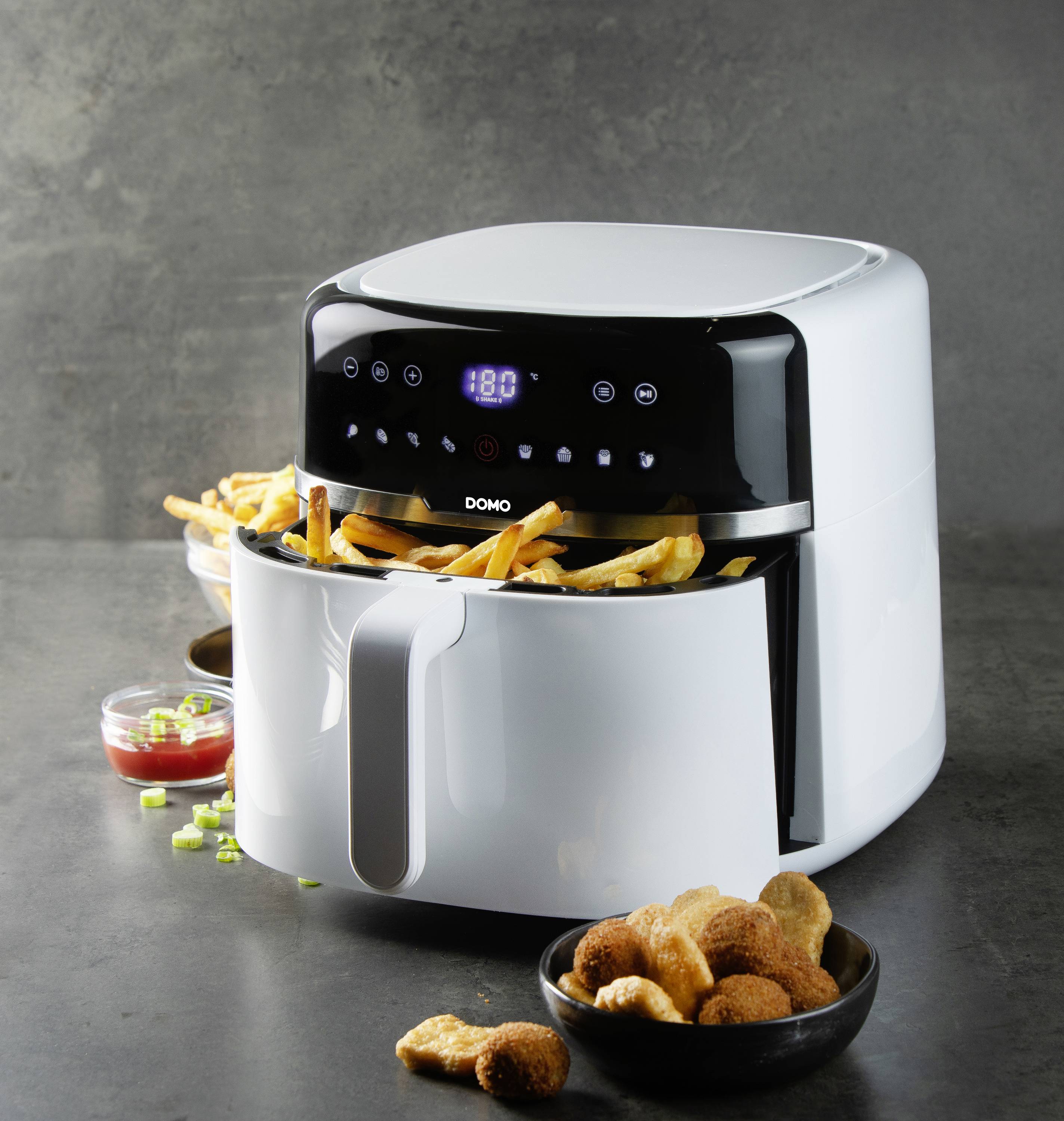 Eine weiße Heißluftfritteuse mit geöffnetem Korb voller Pommes frites. Daneben Schalen mit Dip und Knabbereien auf einer dunklen Oberfläche.