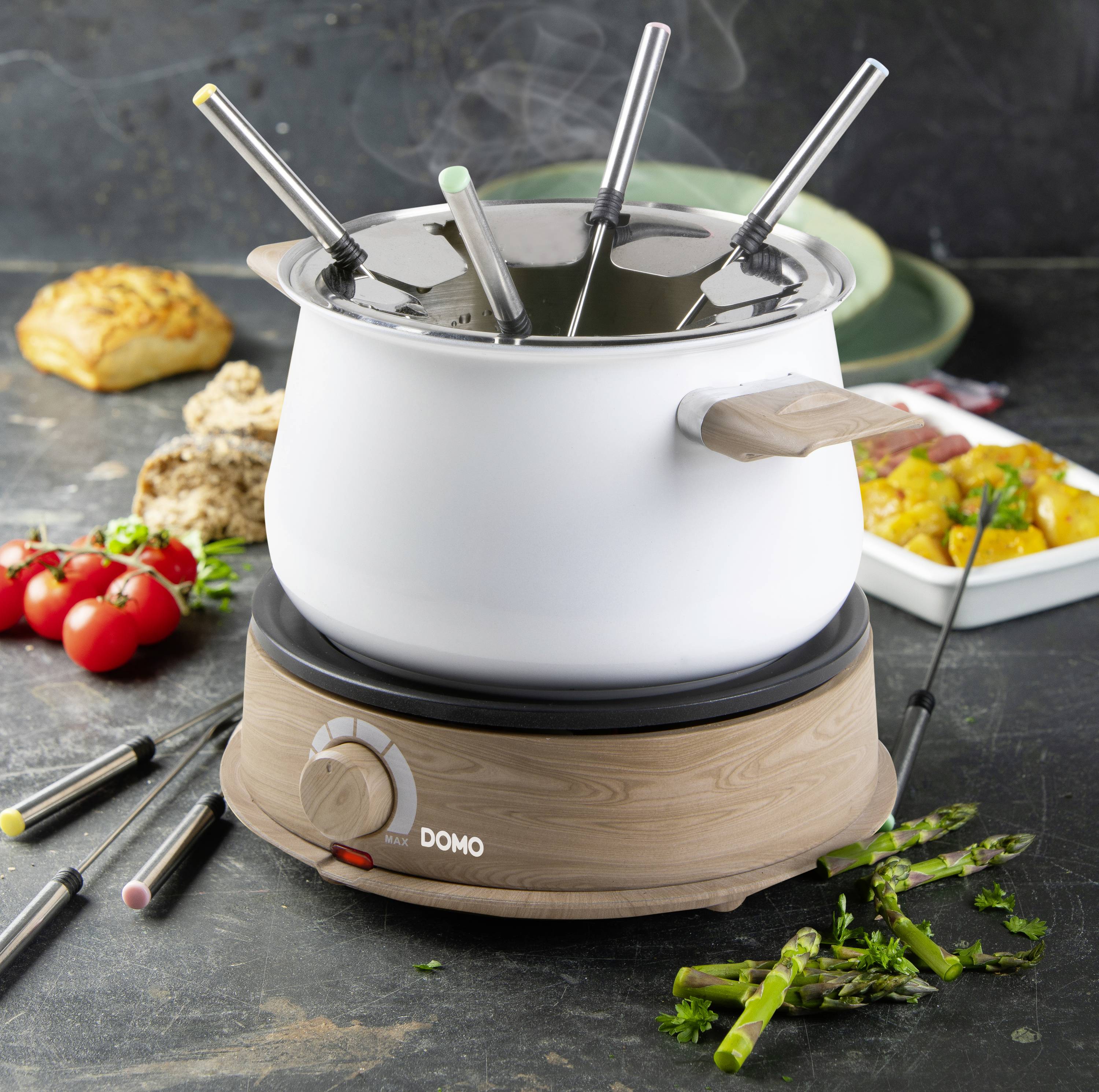 Ein elektrisches Fondue-Set auf einem Tisch, umgeben von Gemüse und Brot. Dampf steigt aus dem Topf auf, vier Fondue-Gabeln sind darin.