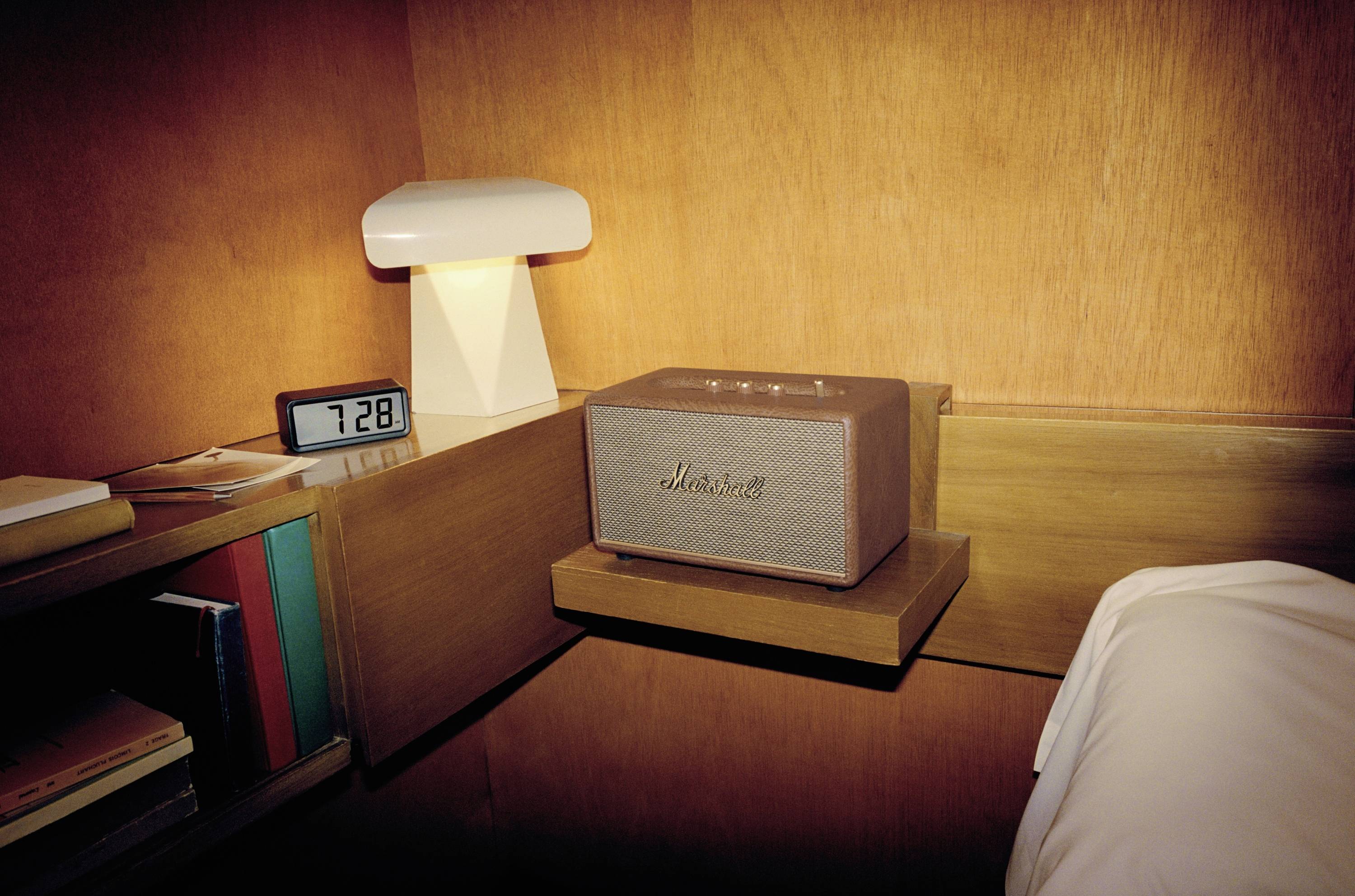 Holzvertäfeltes Schlafzimmer mit Regal, Tischleuchte, Wecker und Lautsprecher. Uhrzeit zeigt 7:28. Regal enthält Bücher.