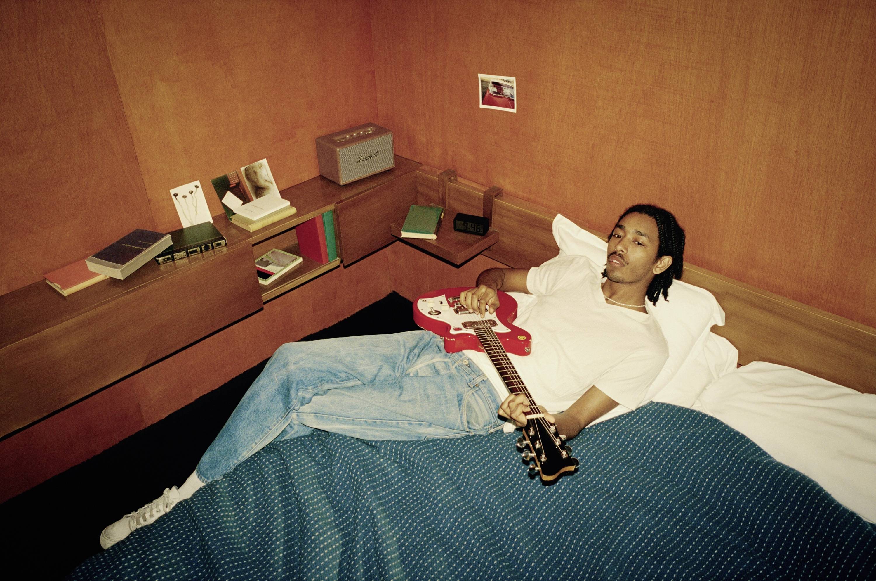 Eine Person liegt auf einem Bett in einem Zimmer mit orangefarbenen Wänden, hält eine rote E-Gitarre und blickt entspannt zur Seite.