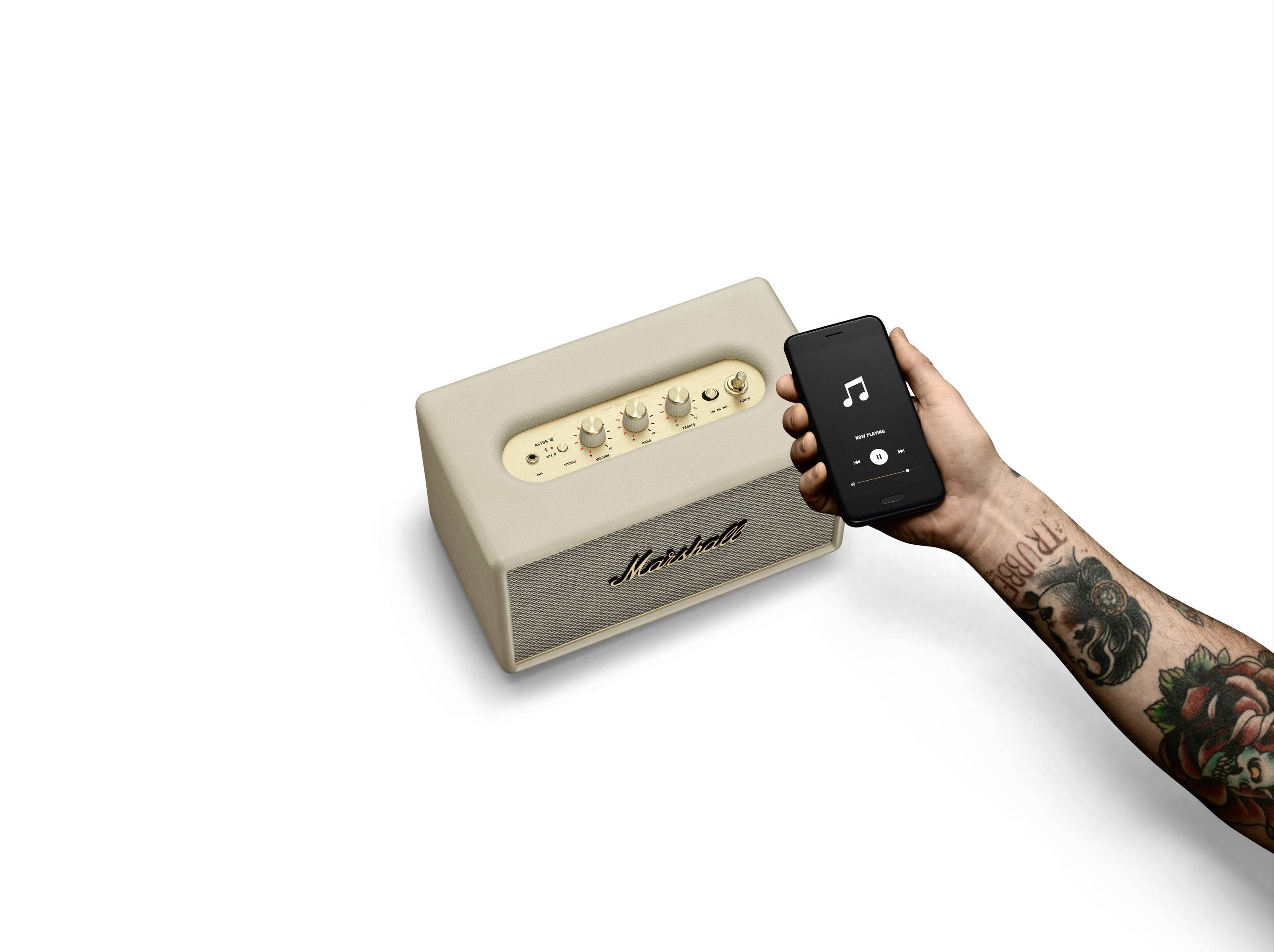 Eine tätowierte Hand hält ein Smartphone mit einer Musik-App neben einem hellen Lautsprecher, der mit 'Marshall' beschriftet ist.