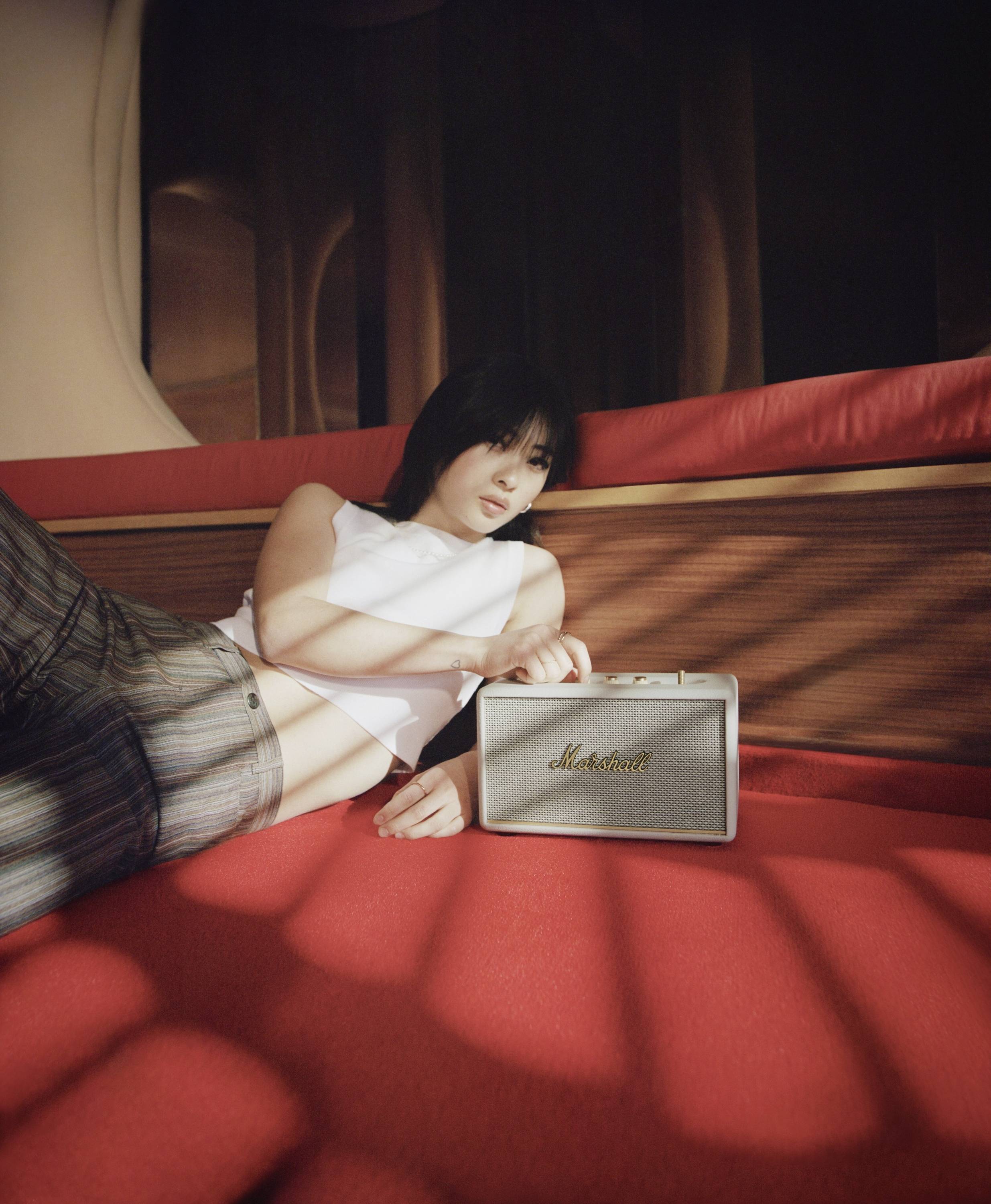 Eine Person liegt entspannt auf einem roten Sofa und hält einen tragbaren Lautsprecher. Sonnenlicht fällt durch ein Fenster hinein.