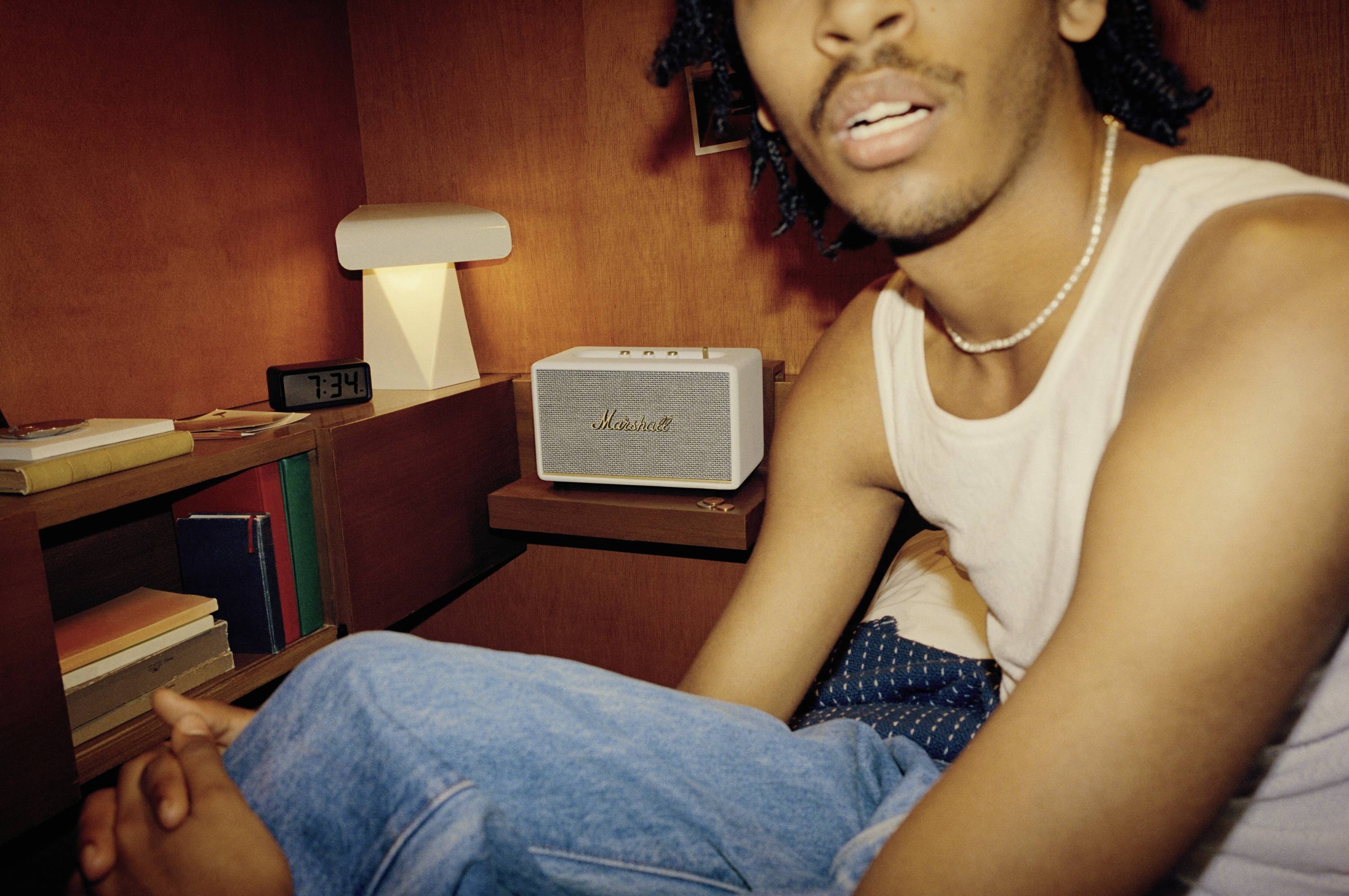 Eine Person sitzt in einem gemütlichen Raum neben einem Lautsprecher, umgeben von Büchern und einer Tischlampe. Es ist 7:34 Uhr.