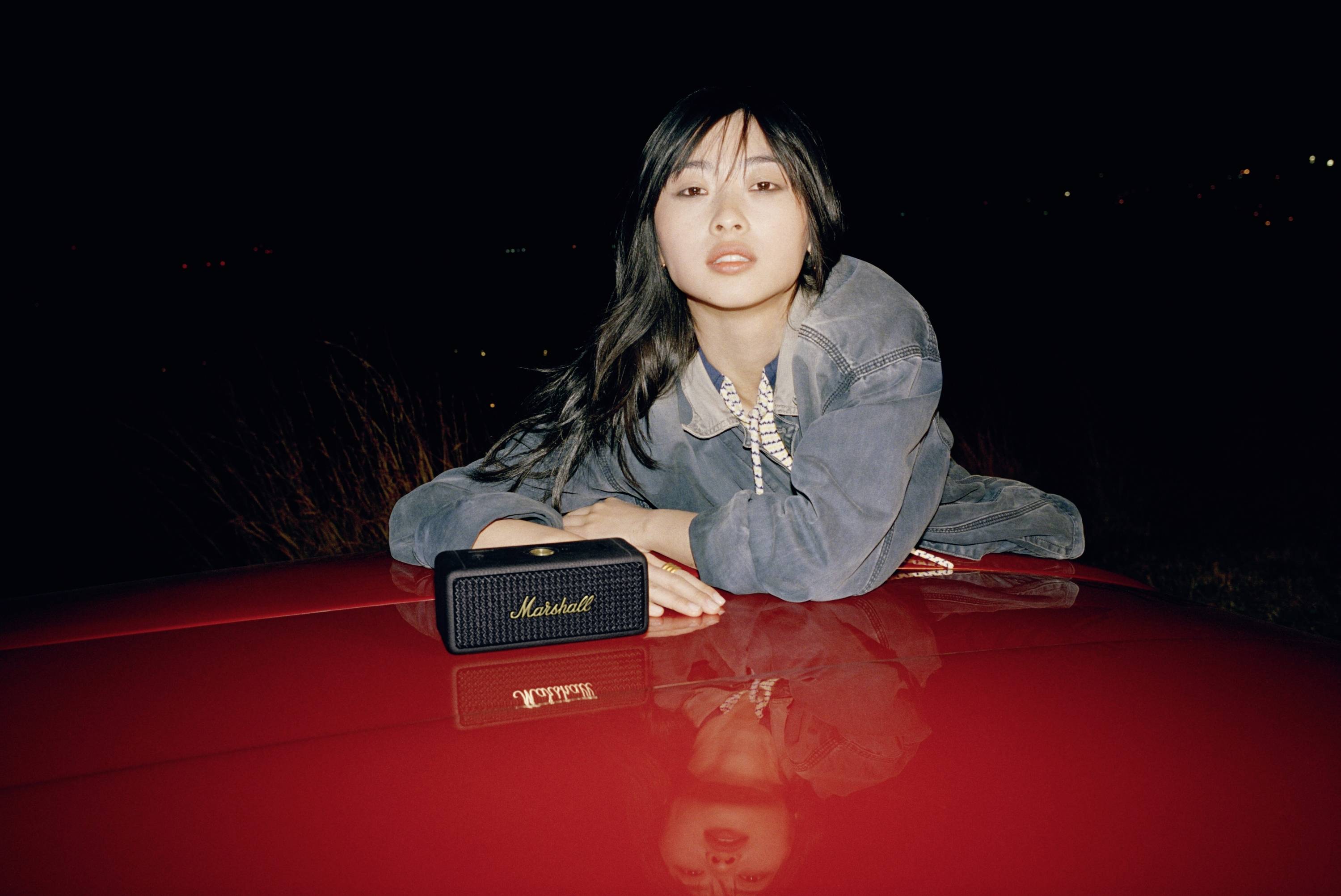 Eine Frau lehnt sich auf ein rotes Auto und schaut in die Kamera, mit einem Lautsprecher vor sich. Es ist Nacht.