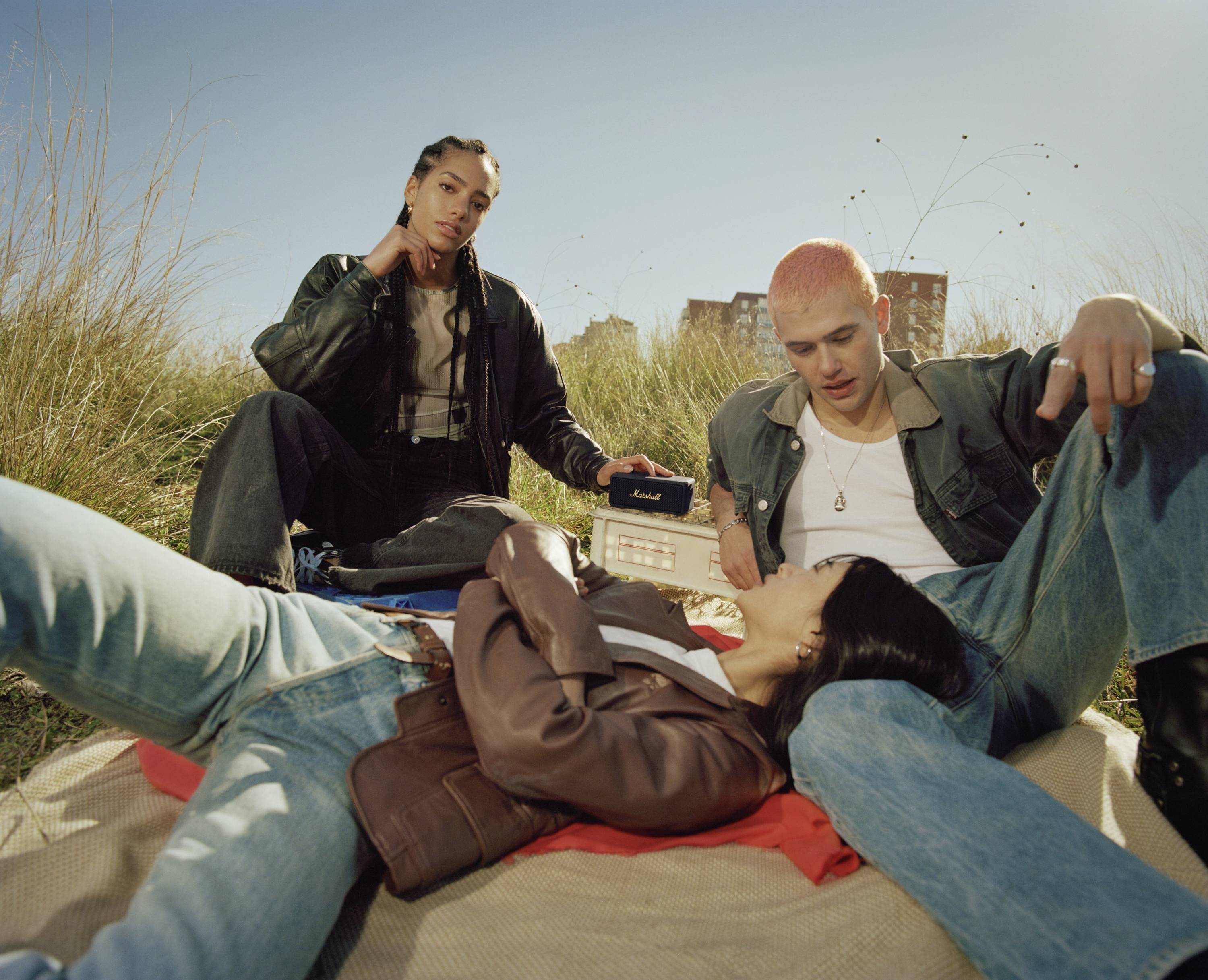 Drei Personen sitzen und liegen auf einer Wiese im Freien. Eine Person hält einen Lautsprecher. Sie wirken entspannt und genießen das Wetter.