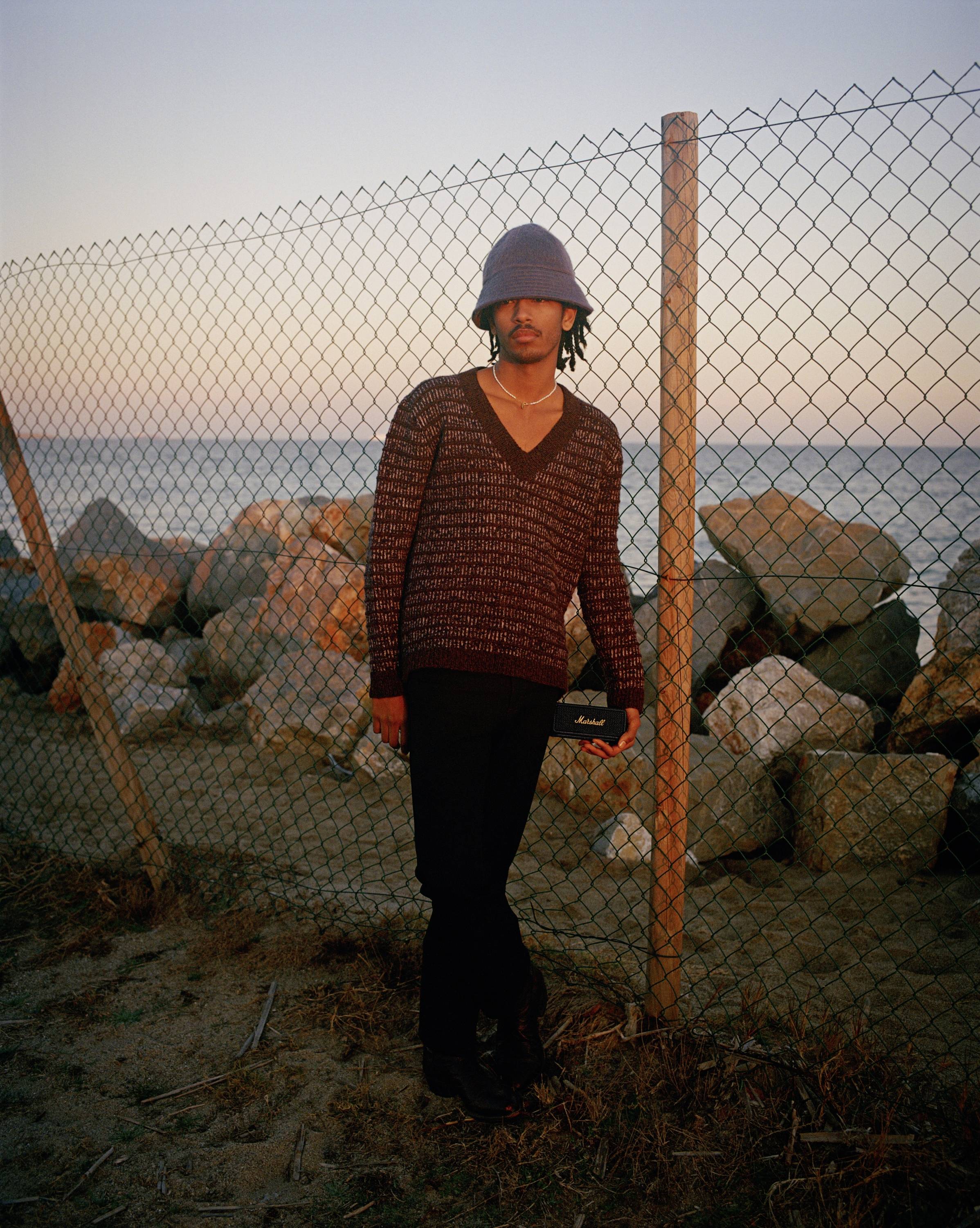Eine Person steht an einem Maschendrahtzaun vor Felsen am Meer bei Sonnenuntergang. Sie trägt einen Hut und hält eine kleine Tasche.