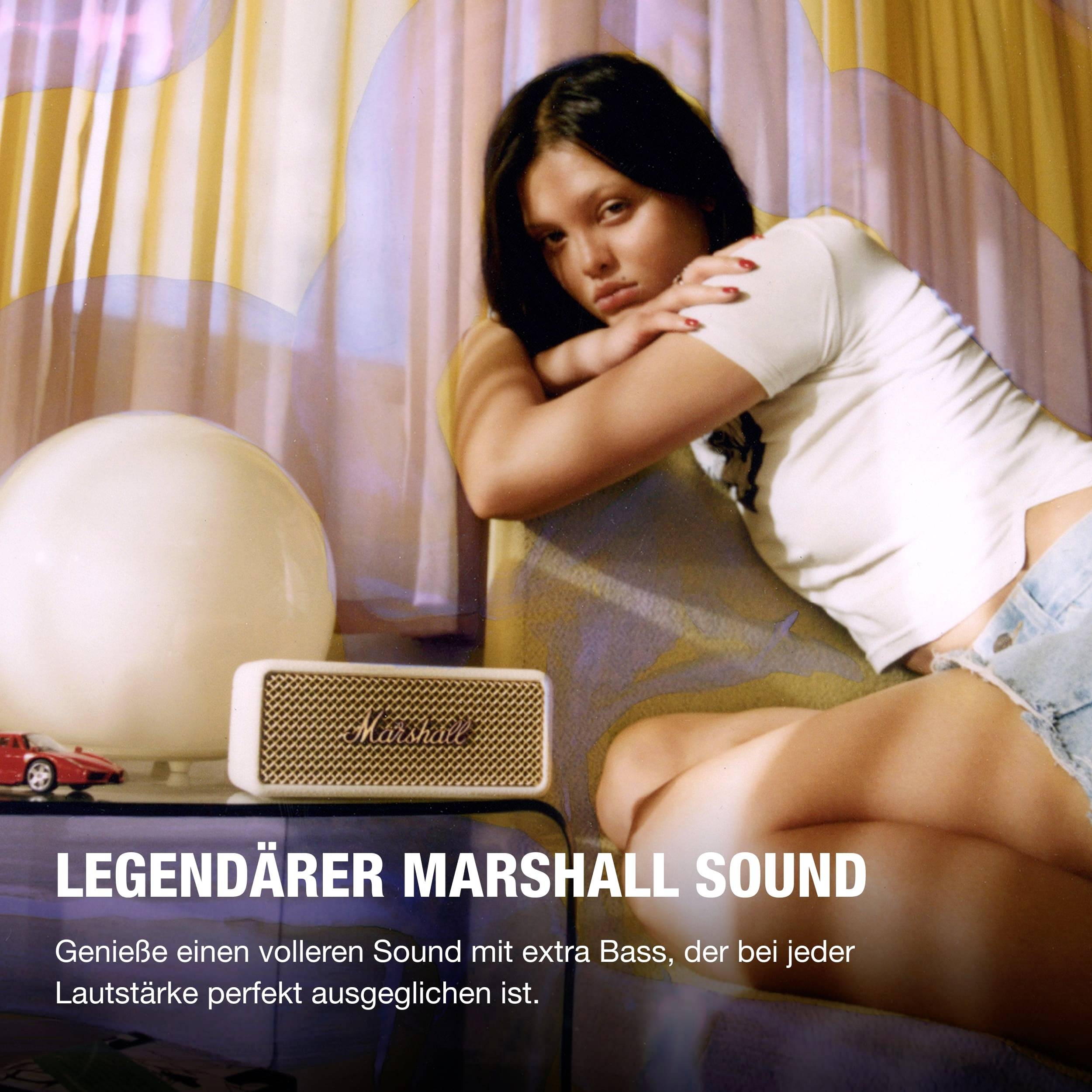 Eine Frau lehnt sich an ein Sofa neben einem Marshall-Lautsprecher. Text: 'Genieße legendären Sound mit extra Bass.'
