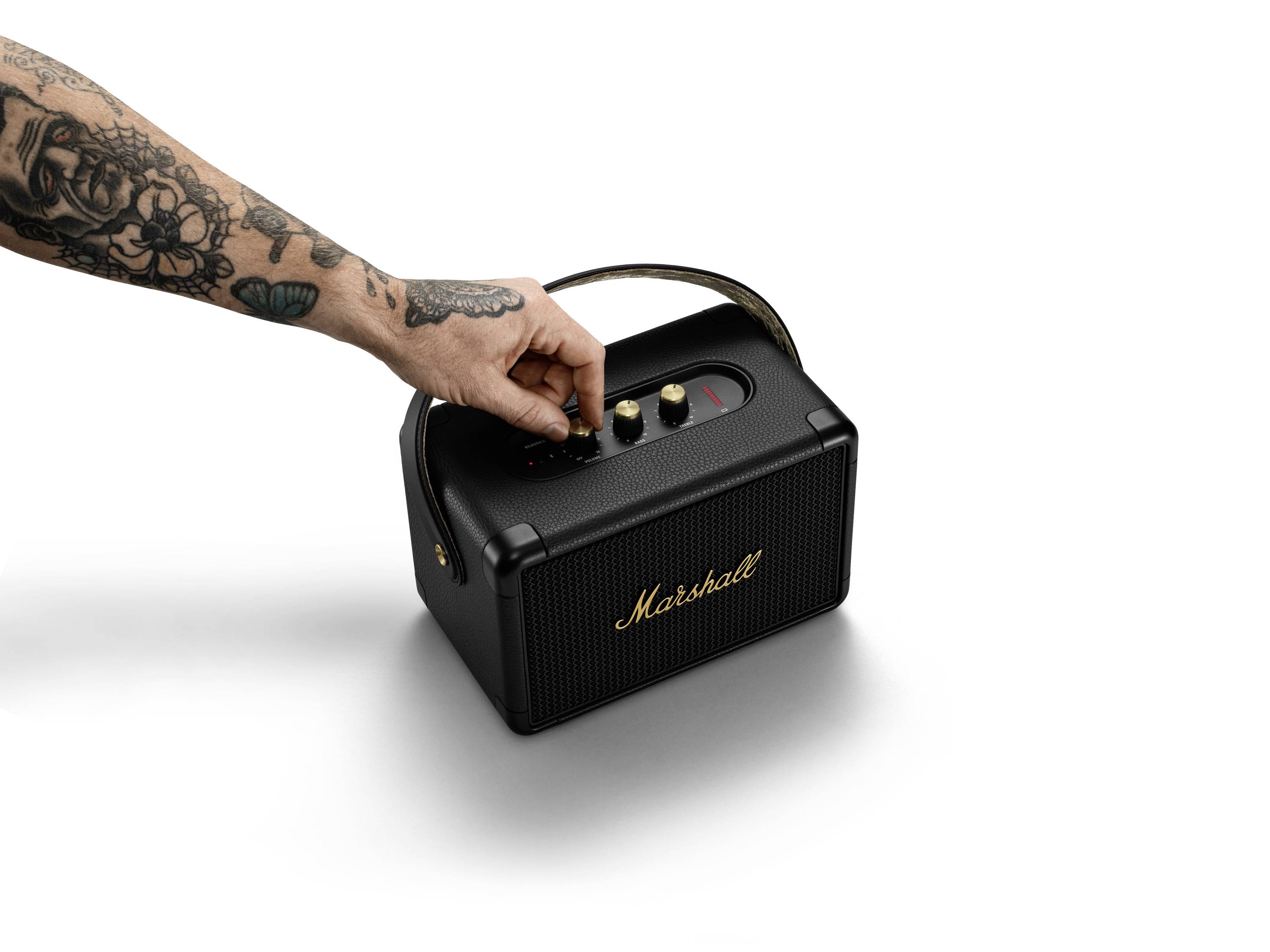 'Tätowierte Hand dreht Knopf auf tragbarem Lautsprecher mit Marshall-Logo auf weißem Hintergrund.'