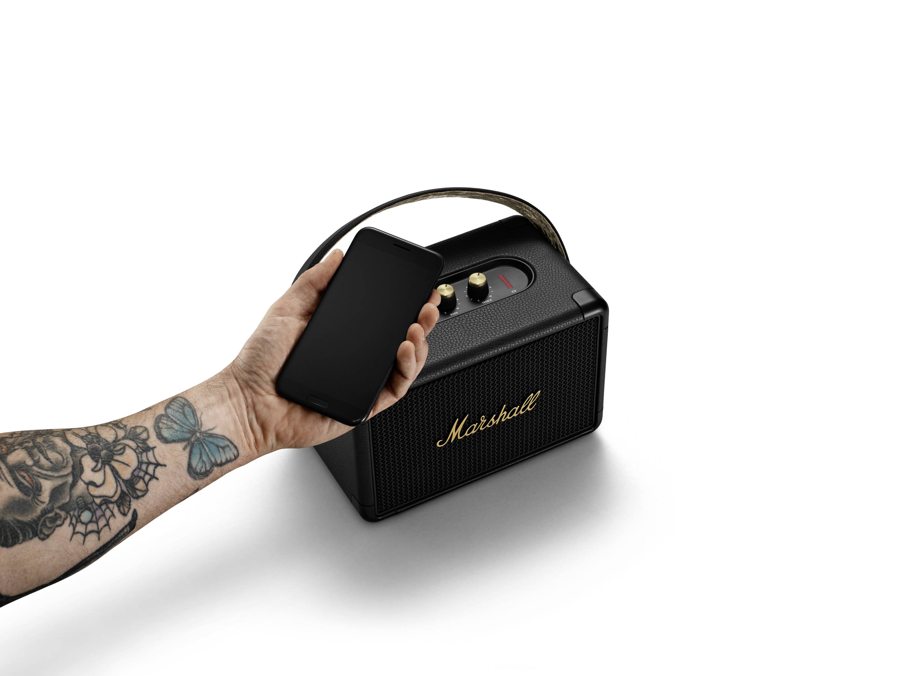 Eine Hand mit Tattoos hält ein Smartphone nahe an einem schwarzen tragbaren Lautsprecher mit goldener Aufschrift auf einem weißen Hintergrund.