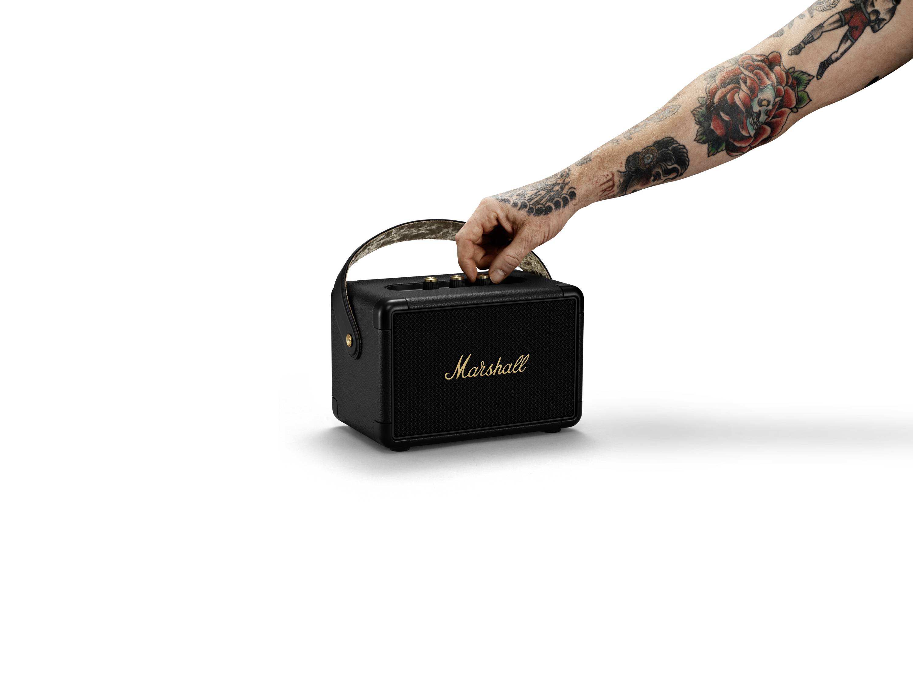 Tragbare schwarze Lautsprecherbox mit einer Aufschrift 'Marshall' und einem Tragegriff. Eine tätowierte Hand dreht den Knopf.