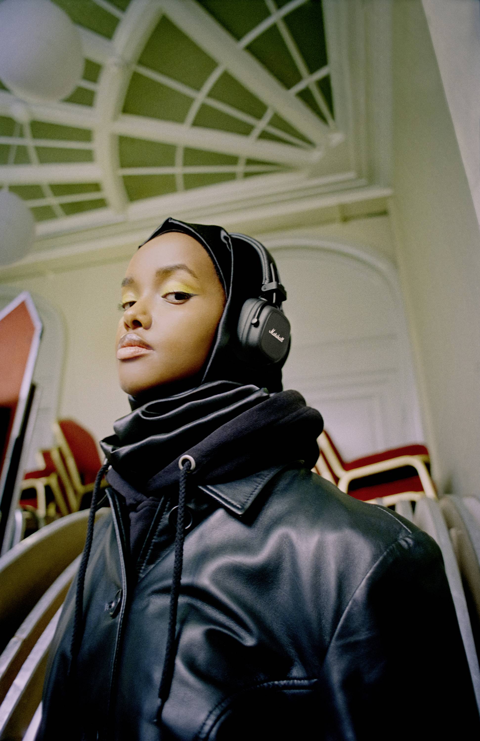 Person in schwarzer Lederjacke und schwarzem Kopfhörer, steht in einem Innenraum mit architektonischen Details und roten Stühlen im Hintergrund.