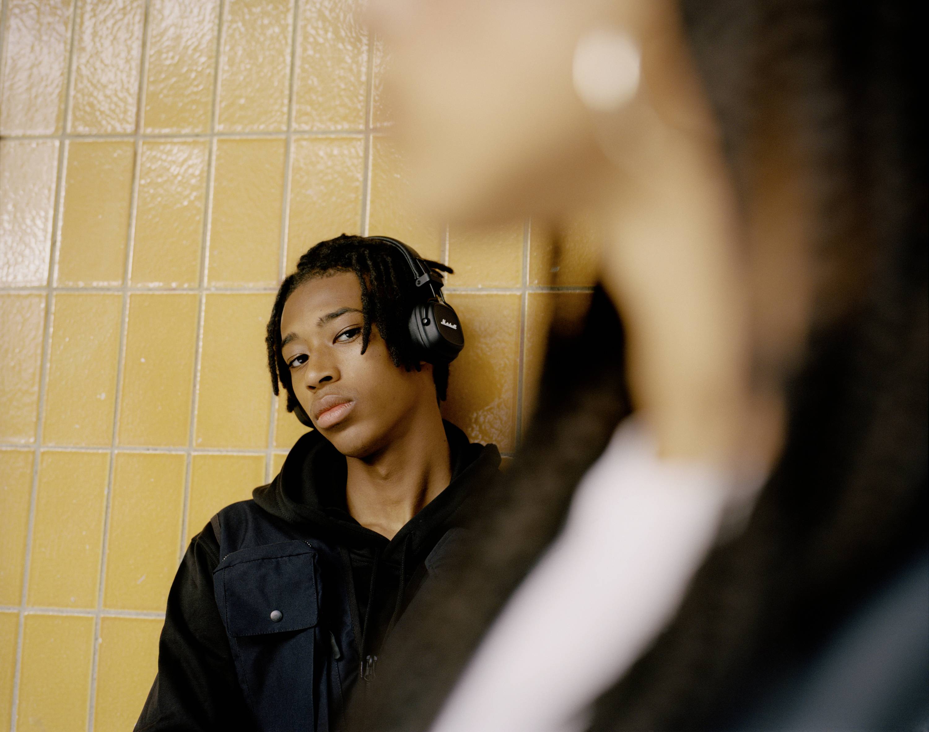 Eine Person mit Kopfhörern lehnt an einer gelben Kachelwand, blickt nachdenklich, während im Vordergrund eine andere Person unscharf ist.