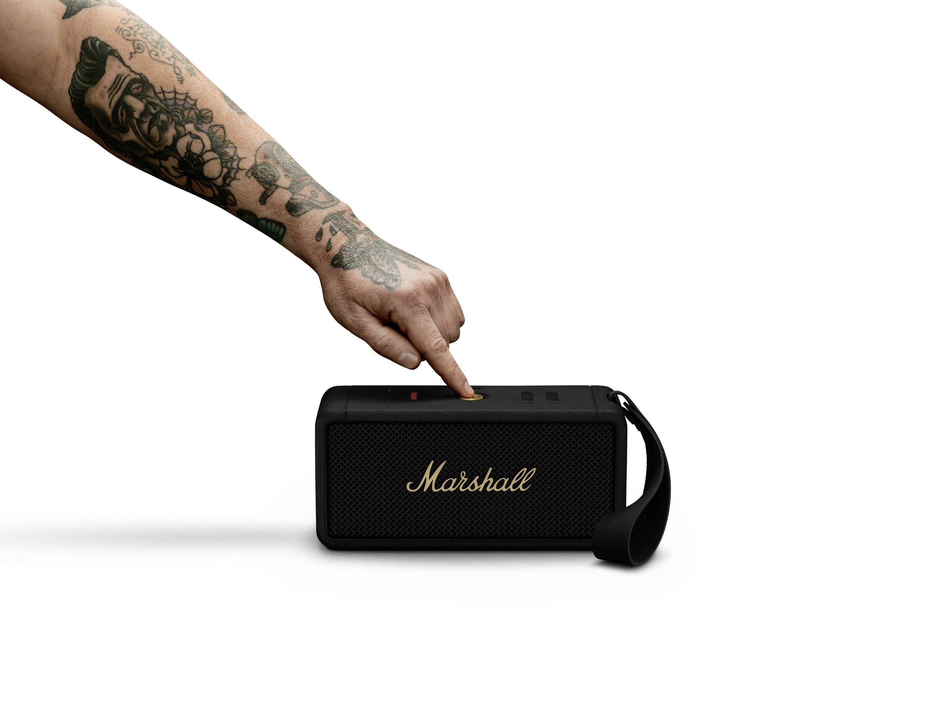 'Eine tätowierte Hand drückt die Taste auf einem schwarzen Marshall-Lautsprecher mit Trageriemen, der auf einer weißen Oberfläche steht.'