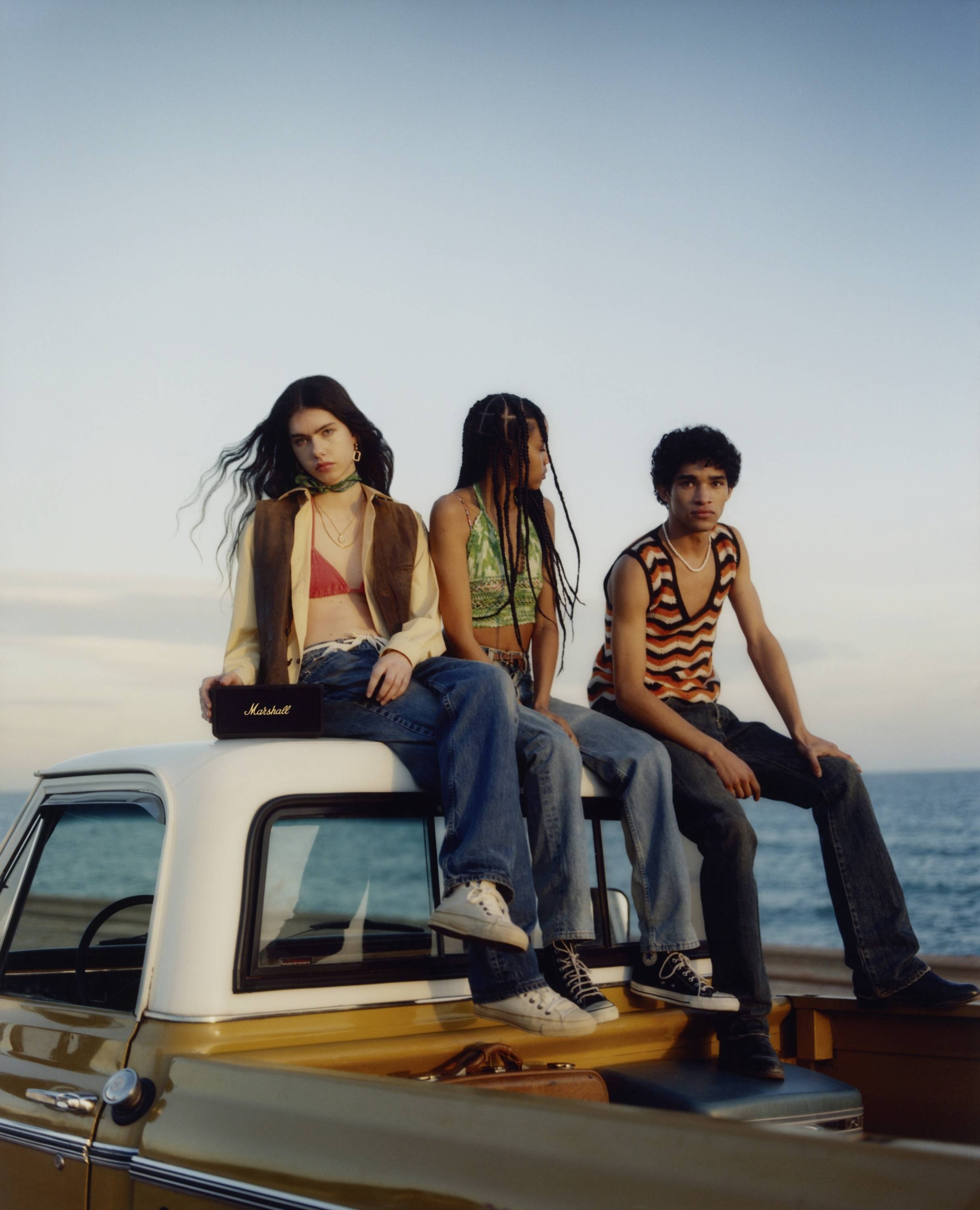 Drei junge Erwachsene sitzen auf einem Auto am Meer bei Sonnenuntergang, lässig gekleidet, der Hintergrund zeigt das ruhige Wasser.