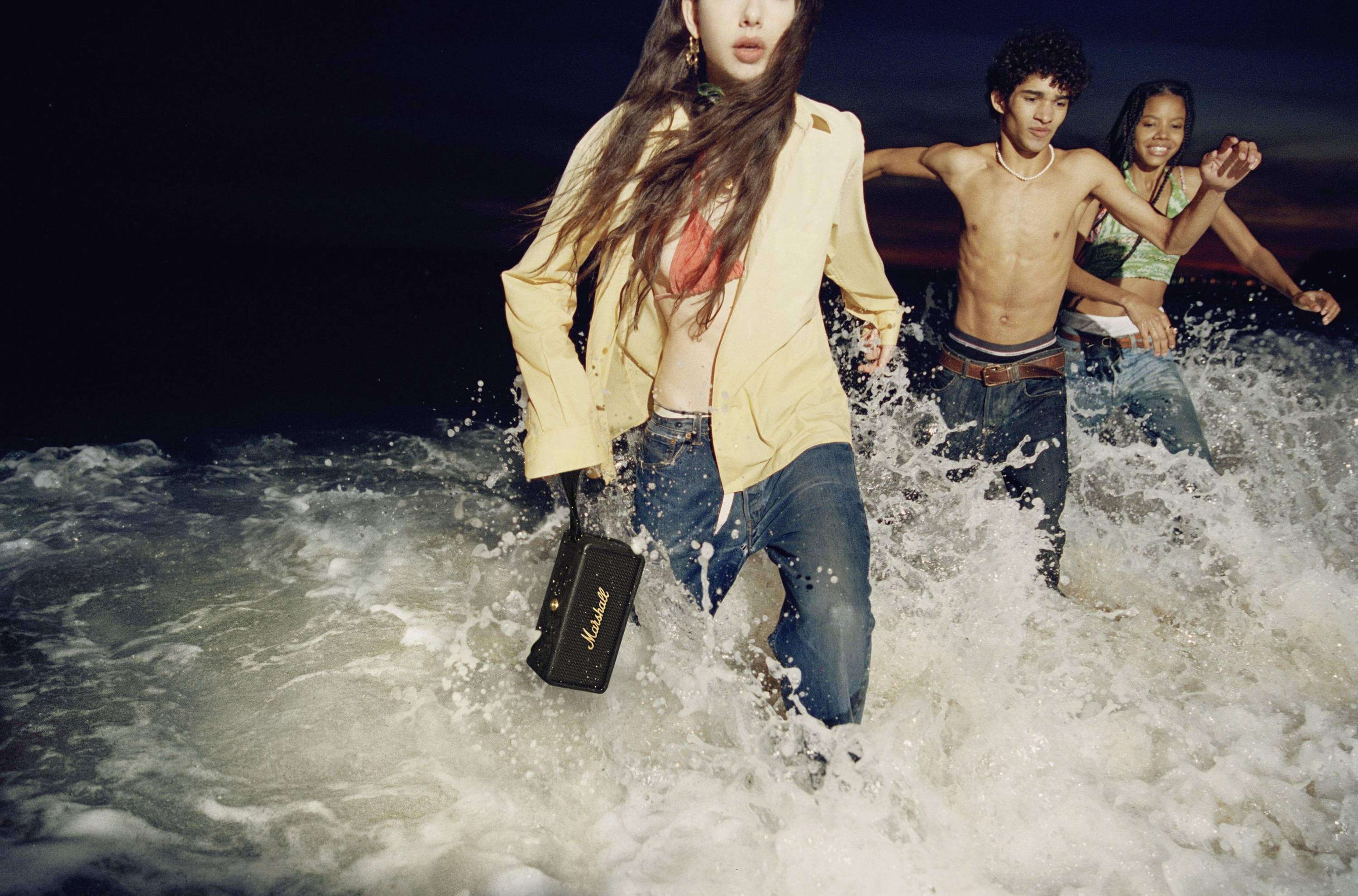 'Drei Personen rennen lachend durch spritzende Wellen am Strand. Eine Person trägt eine gelbe Jacke und hält eine schwarze Tasche.'