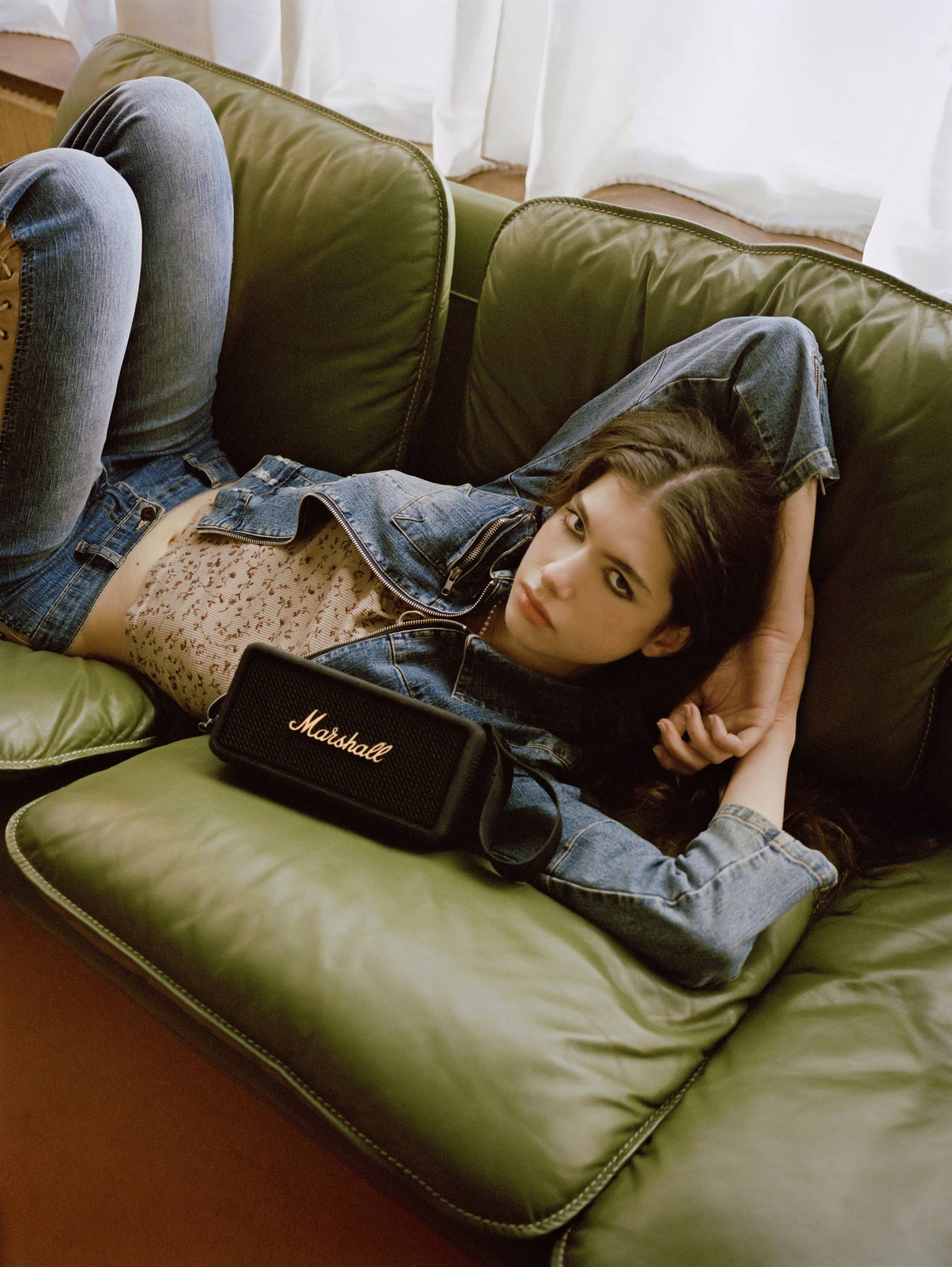Frau liegt entspannt auf einem grünen Sofa und hält eine kleine schwarze Marshall-Lautsprechertasche, trägt Jeans und eine Jeansjacke.