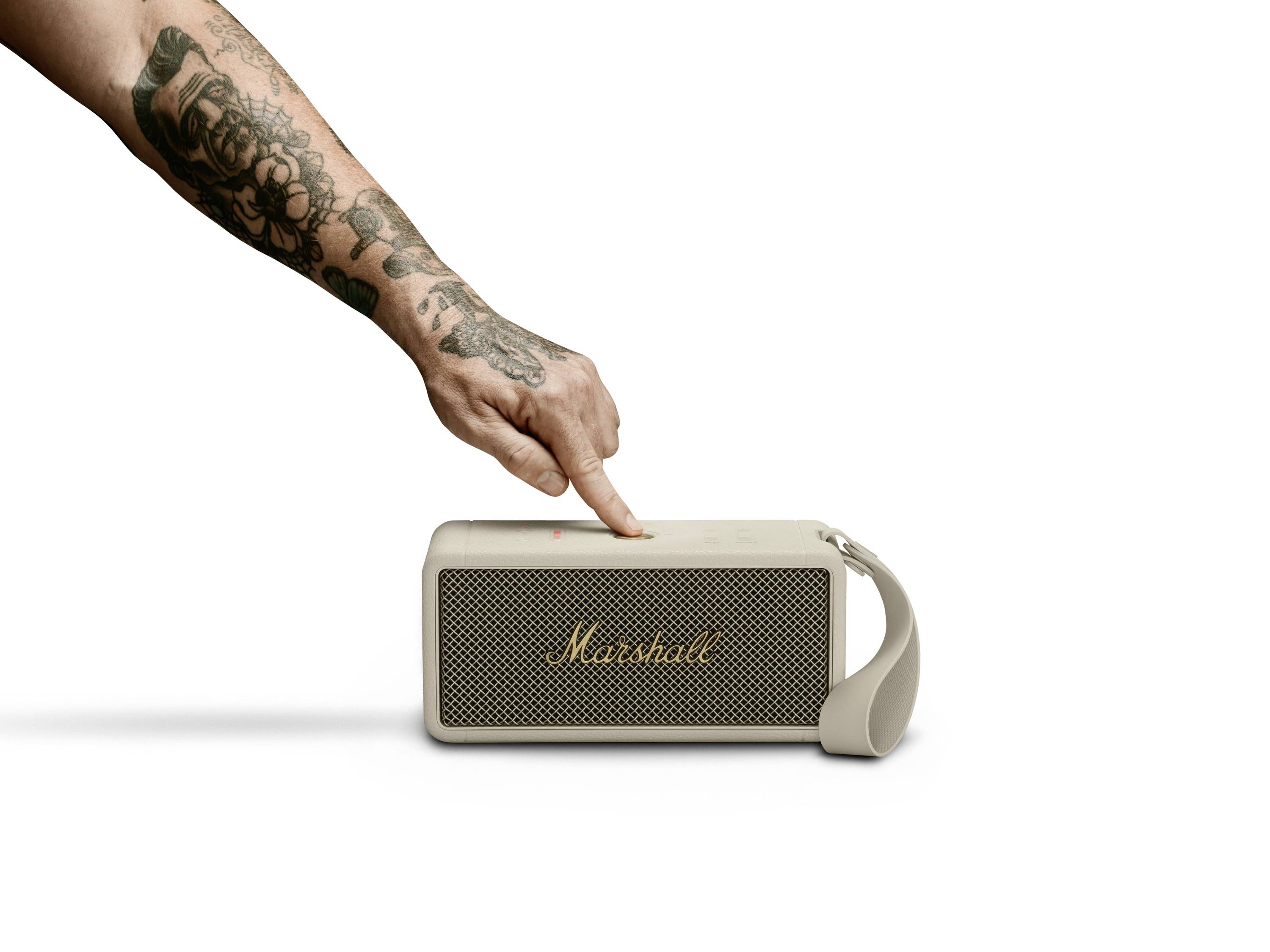Eine tätowierte Hand drückt auf den Lautstärkeregler eines tragbaren Lautsprechers der Marke Marshall, der auf einem Tisch liegt.