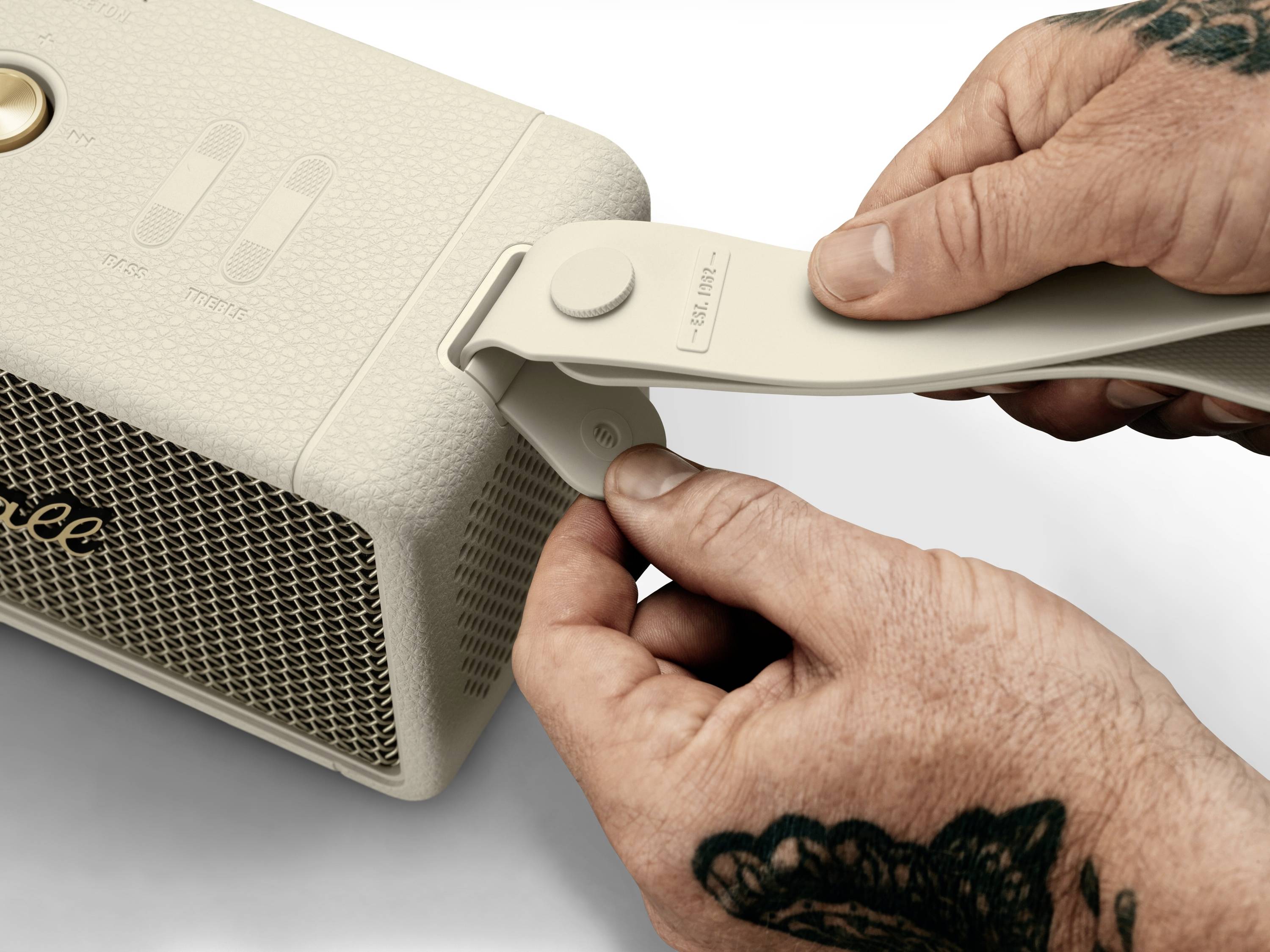 Eine Person befestigt einen Tragegurt an einem tragbaren Lautsprecher. Der Lautsprecher hat ein Textur-Design und seitliche Bedienelemente.