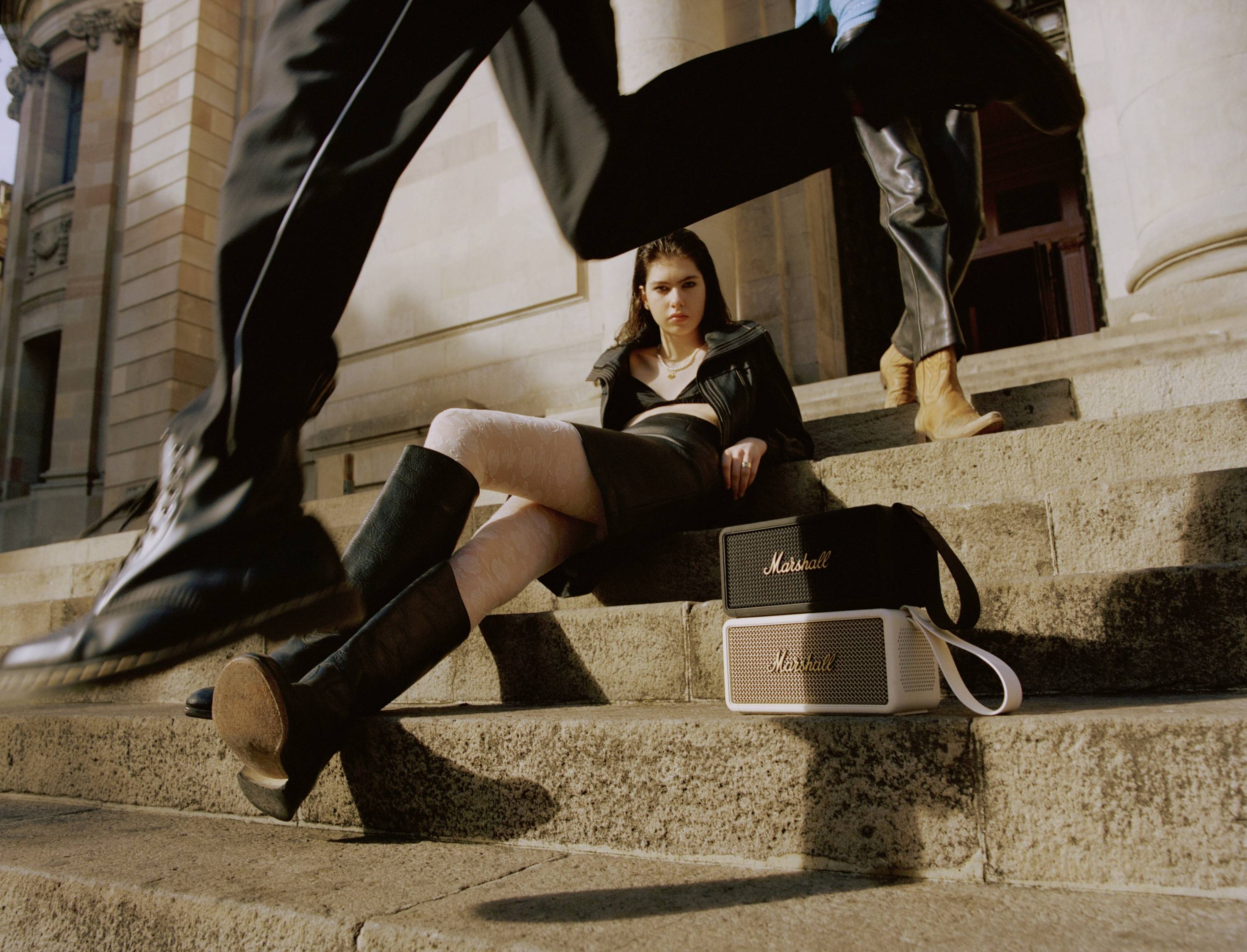 Person sitzt auf Treppe mit zwei Lautsprechern der Marke Marshall. Eine Person in Bewegung läuft vorbei. Gebäude mit Säulen im Hintergrund.