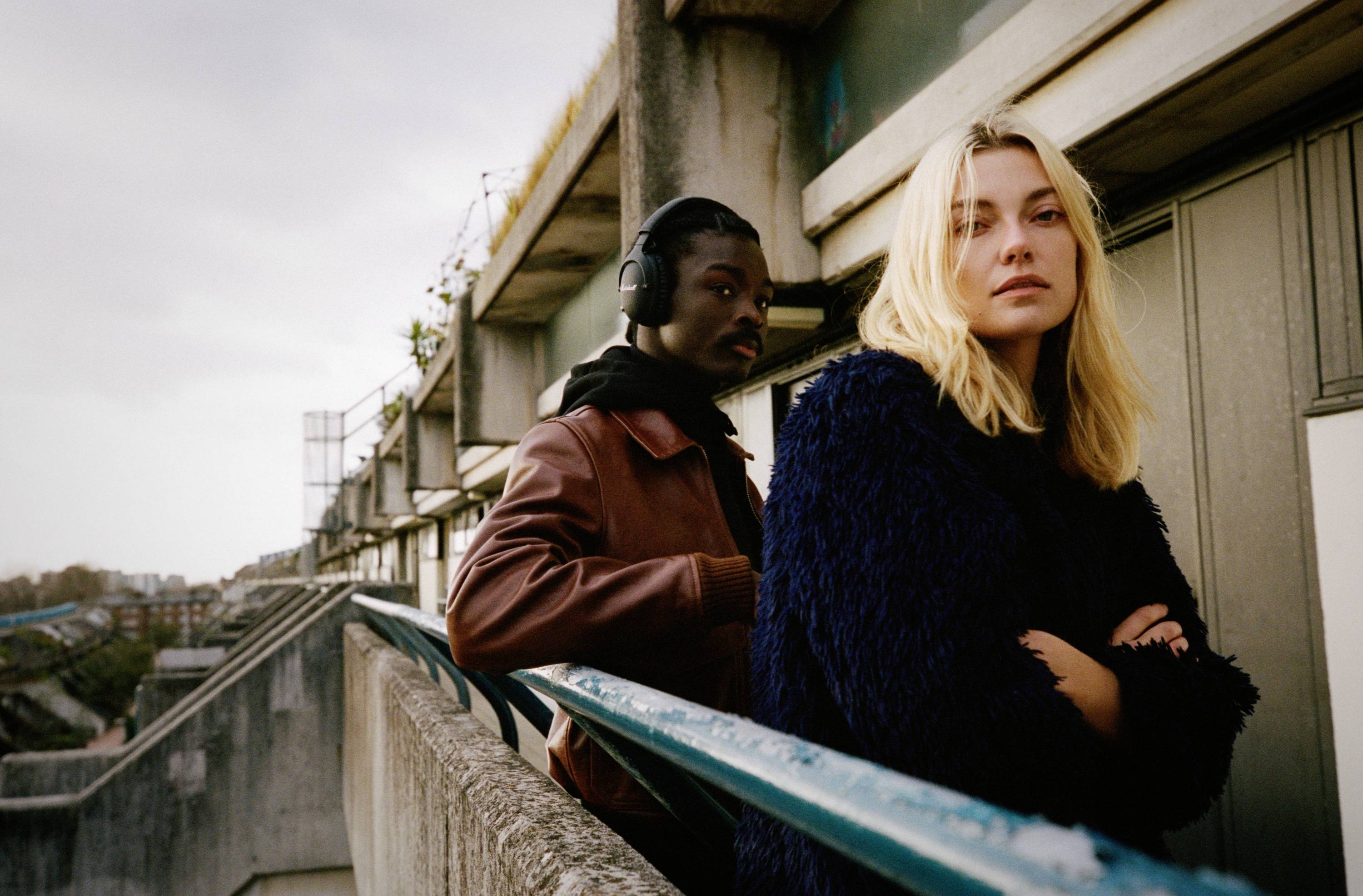 Zwei Personen stehen auf einem Balkon, draußen. Eine Person mit Kopfhörern schaut nach unten, die andere schaut direkt in die Kamera.